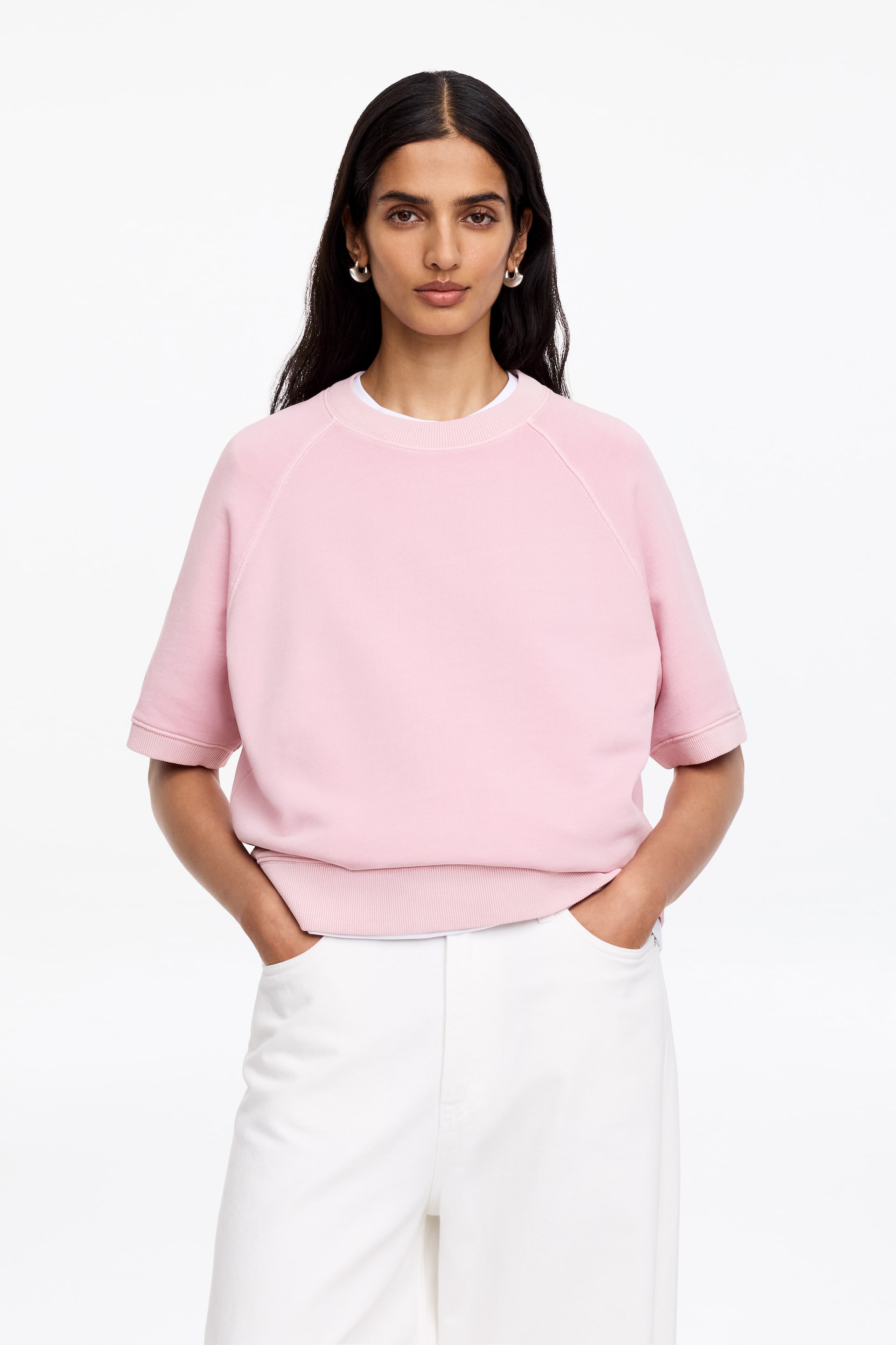 View larger image: The model is wearing a pink short-sleeved sweatshirt with a crew neck, raglan-cut sleeves, and rib-knitted trimmings on the collar, cuffs, and hem. It is layered over a white t-shirt and styled with white trousers.