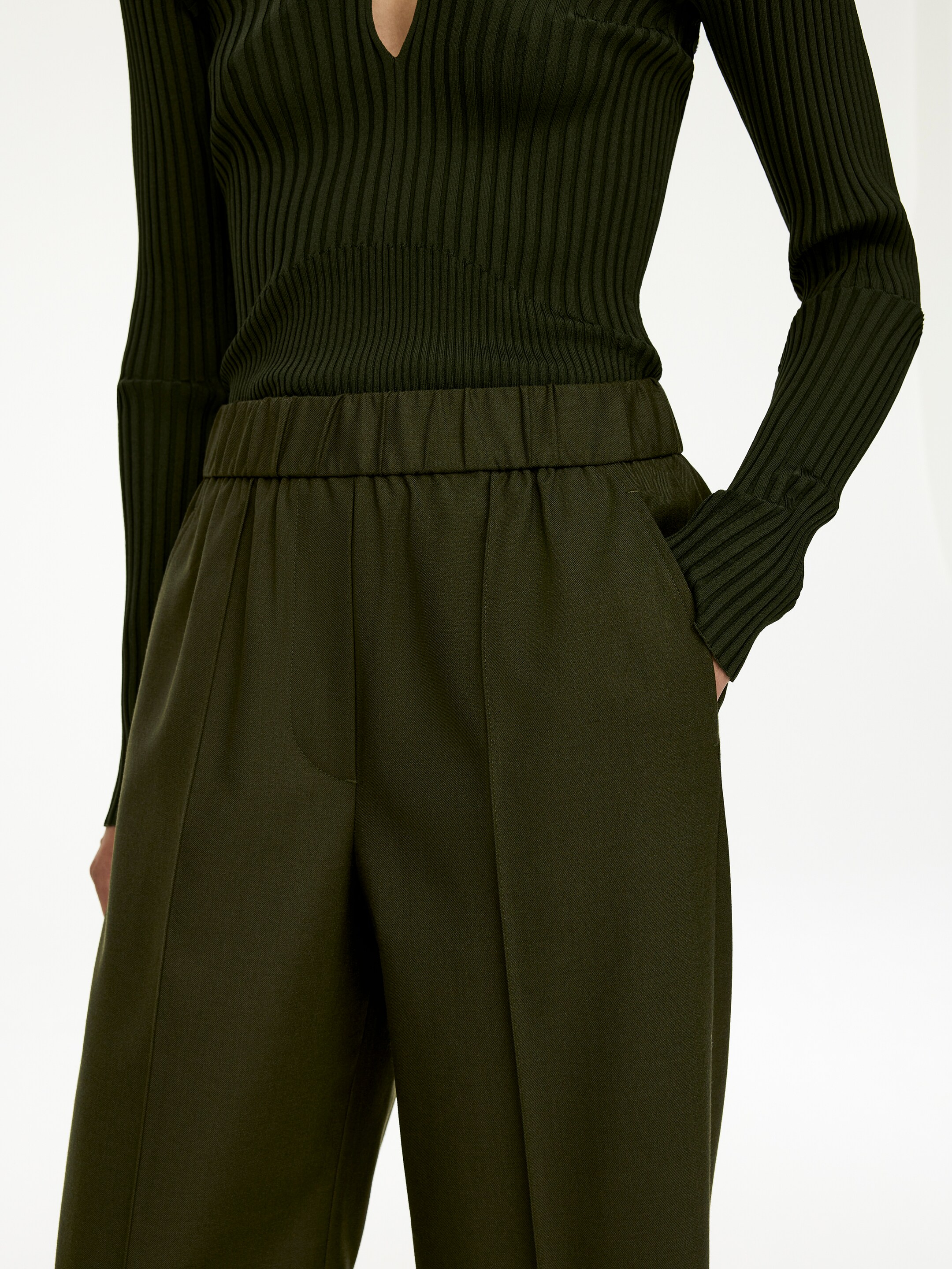View larger image: A dark green ribbed knit top and matching dark green pants are being worn, viewed from a slight front-side angle. The top is featuring a V-neck and long sleeves with flared ribbed cuffs, while the pants are showcasing an elasticated waistband, front leg creases, and side pockets, with a hand resting in one.