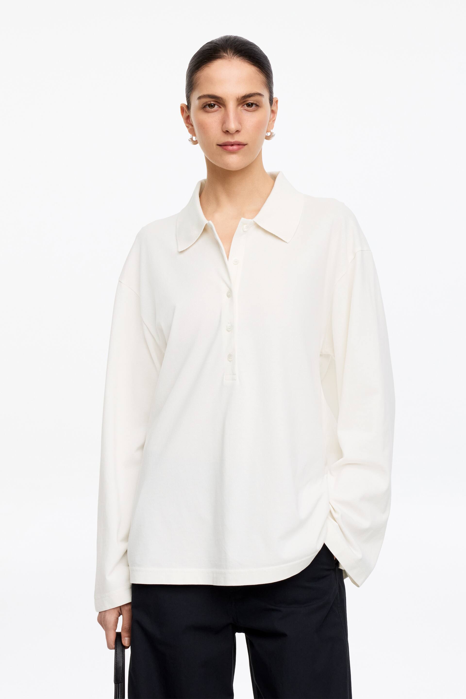 Größeres Bild anzeigen: Ein cremefarbenes Langarm-Poloshirt wird von vorne präsentiert, mit einem spitzen Kragen und einer Knopfleiste mit vier sichtbaren Knöpfen. Das Shirt ist mit einer lockeren Passform und langen Ärmeln gestaltet, und sein gerader Saum liegt über dunklen Hosen.