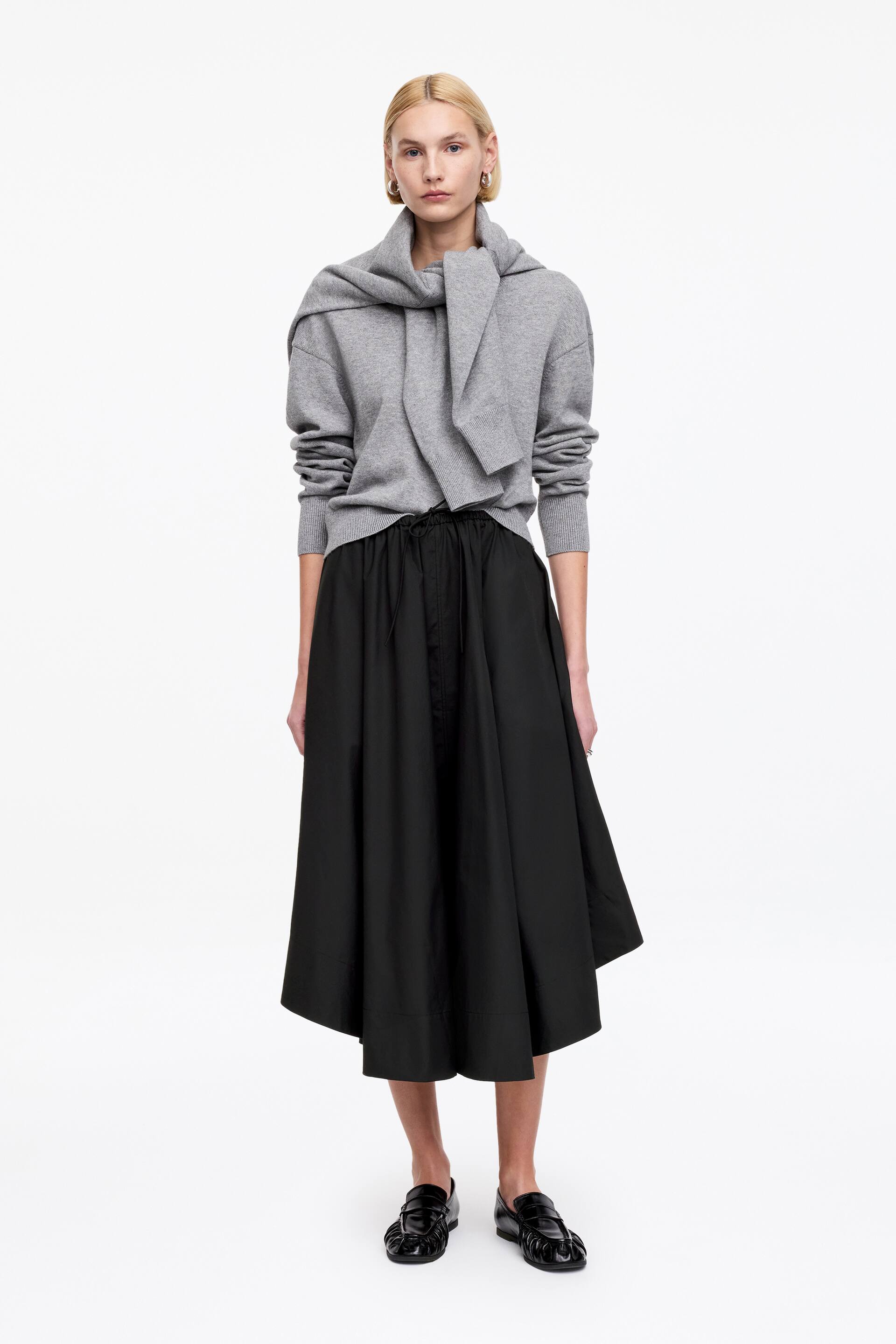 Ver imagen más grande: Un jersey gris de manga larga presenta puños acanalados y un dobladillo acanalado, con una bufanda gris a juego colocada alrededor de su cuello. El jersey está metido en una falda negra de largo midi, que muestra una cintura con cordón y una silueta completa, y se combina con mocasines negros que exhiben un empeine texturizado.