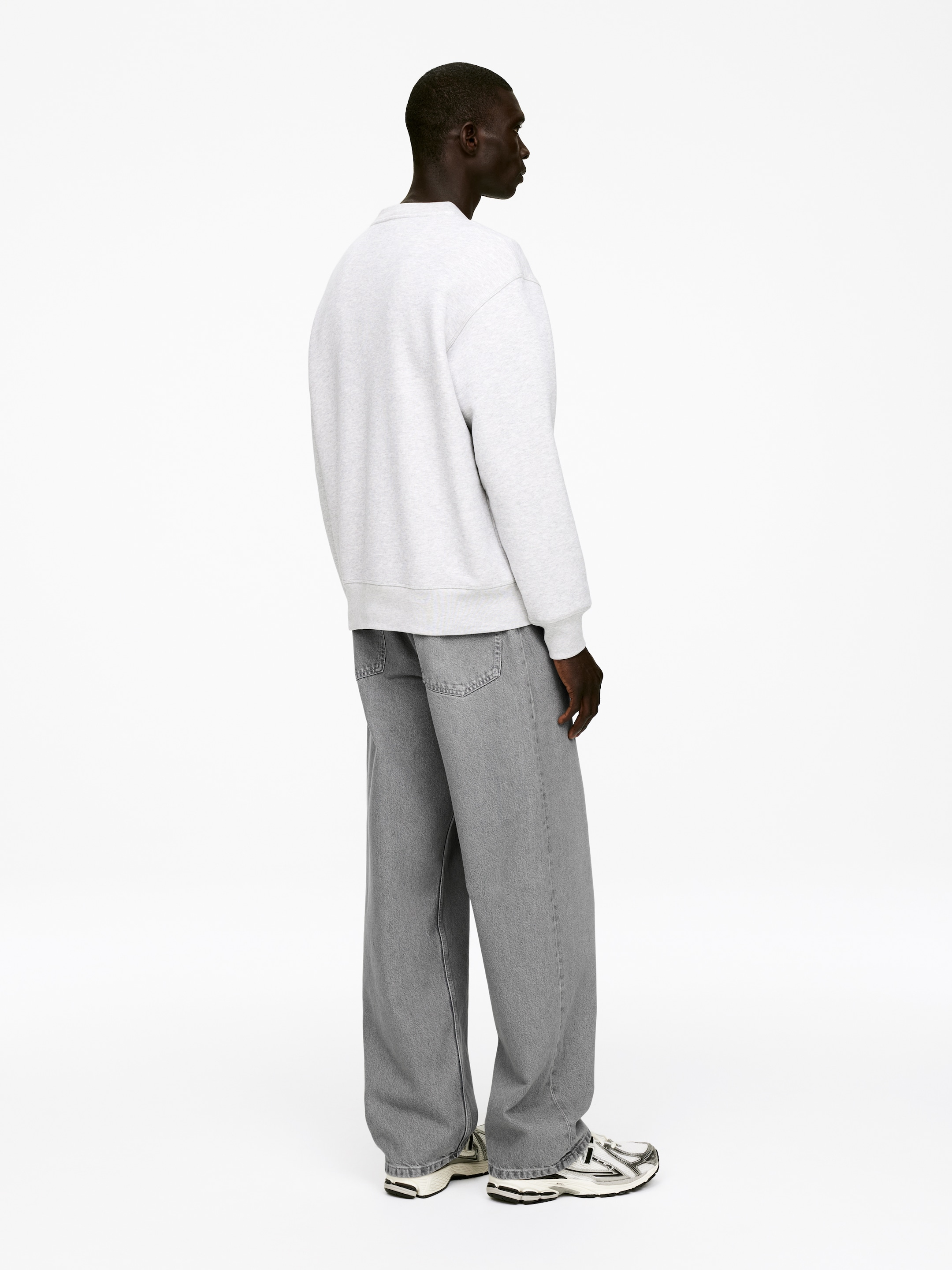 Ingrandisci l'immagine: Una felpa oversize con scollo rotondo in grigio chiaro melange, con polsini e orlo a coste, è indossata da una persona vista da dietro e di lato destro, mostrando la sua vestibilità rilassata. Jeans grigi a gamba larga e vestibilità ampia coprono le gambe, sovrapponendosi a scarpe da ginnastica bianche e nere.