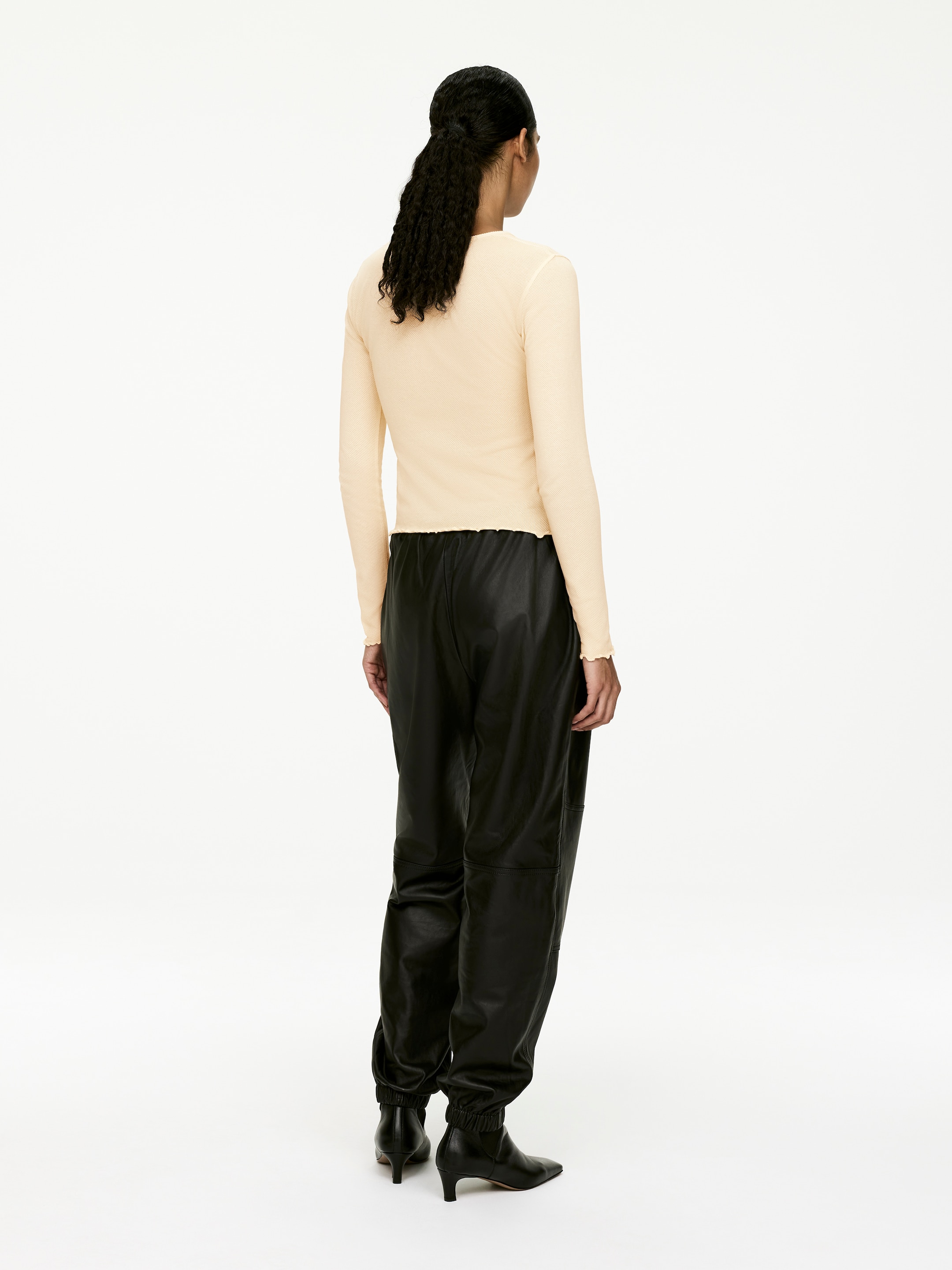 Agrandir l'image: Le haut beige clair à manches longues est présenté de dos, avec une coupe ajustée et des ourlets et poignets bordés de volants. Il est porté avec un pantalon fuselé aspect cuir noir avec des coutures visibles et des poignets élastiqués froncés, qui se terminent au-dessus des bottines noires à bout pointu.