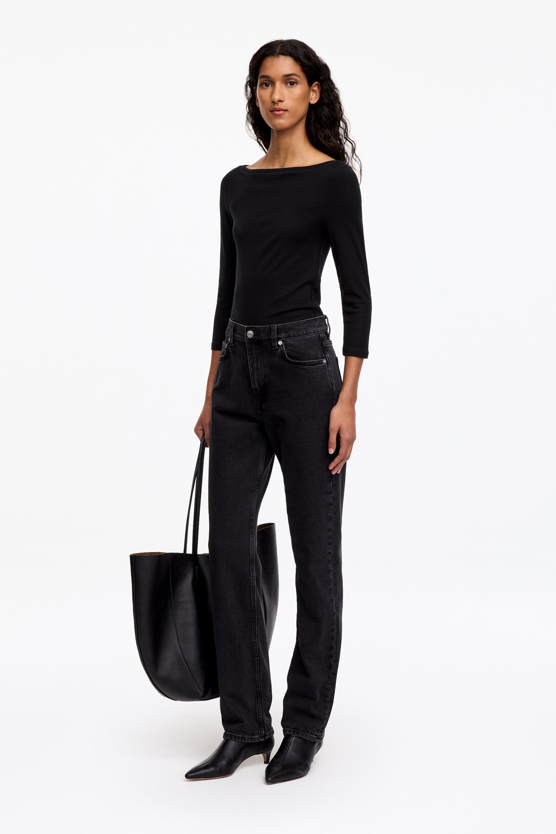 Visualizza l'immagine ingrandita: Un top nero slim fit con scollo a barca e maniche a tre quarti è indossato dalla modella, infilato in un paio di jeans neri a gamba dritta. L’outfit è completato da una grande tote bag nera in pelle tenuta dalla modella e da stivali neri con tacco e punta affusolata.