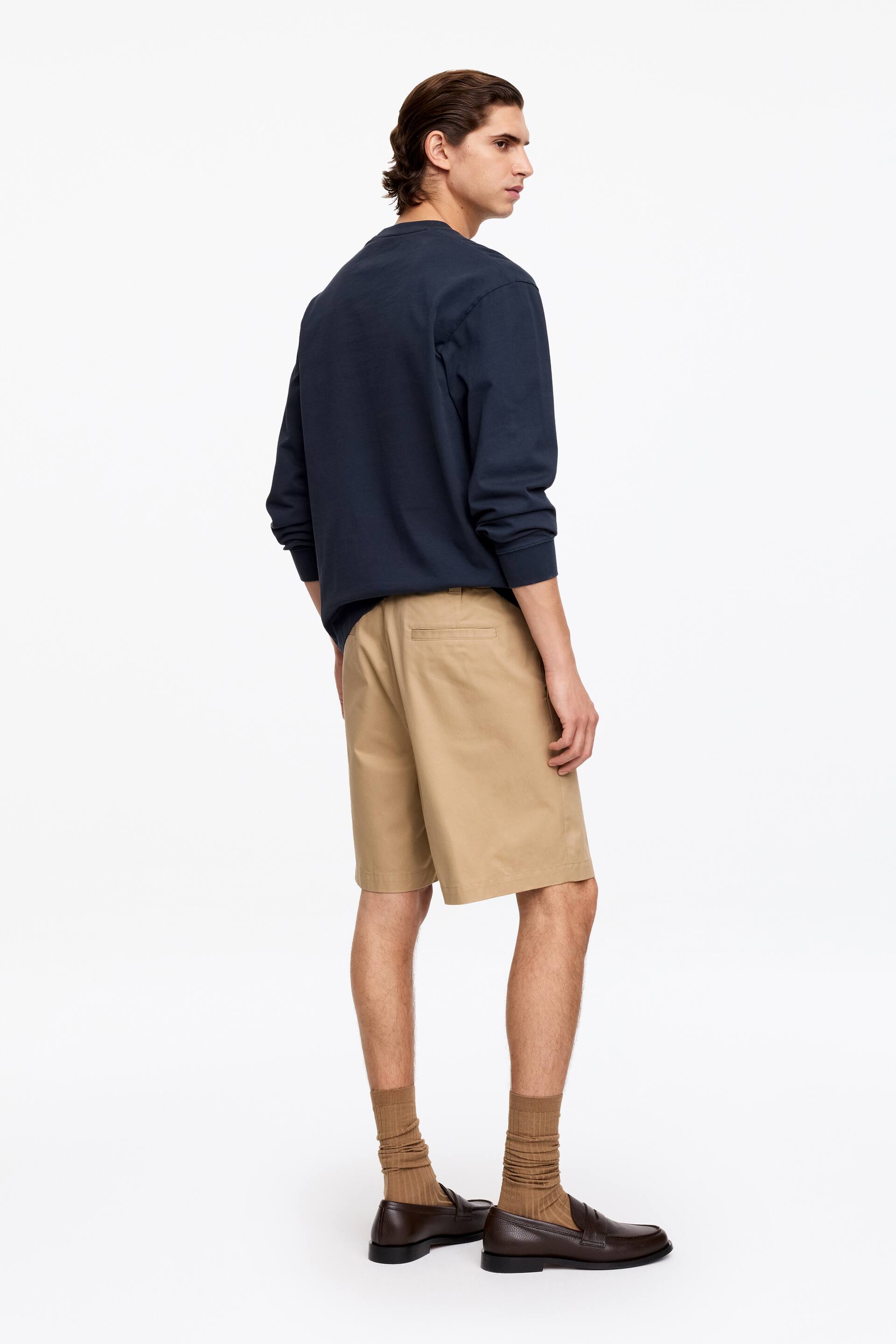 Visa större bild: En marinblå långärmad tröja med rund hals bärs tillsammans med khaki chino shorts, presenterad på en person sedd från bakifrån och höger sida. Shortsen når precis ovanför knäna, och mörkbruna loafers bärs med ribbade bruna strumpor.