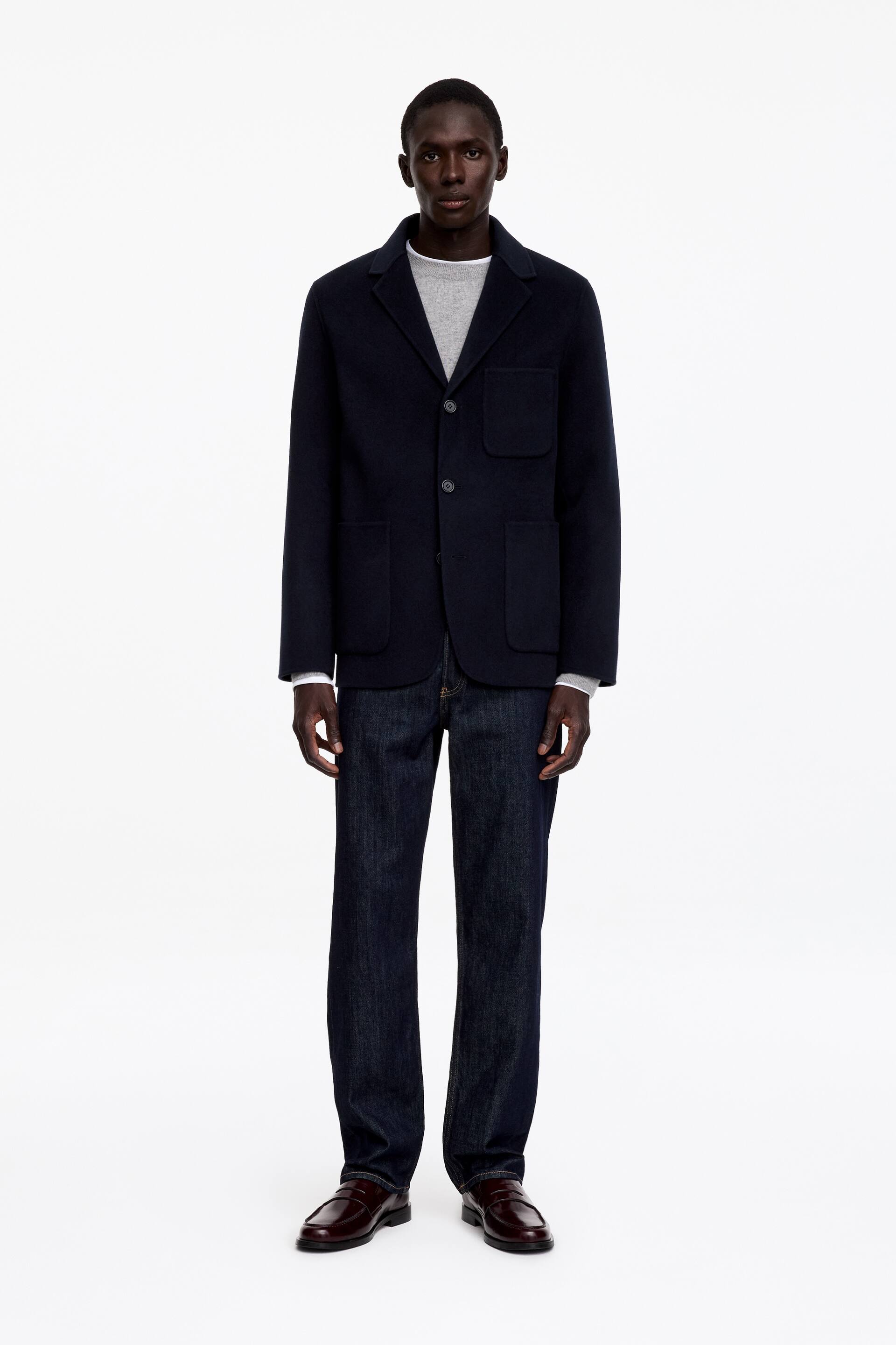 Visa större bild: Modellen bär en mörkblå blazer med klassiska slag, tre knappar framtill med en knapp knäppt, en bröstficka med påsydd kant samt två lägre påsydda fickor. Modellen står upp och bär blazern över en ljusgrå tröja, mörka jeans och bruna loafers.
