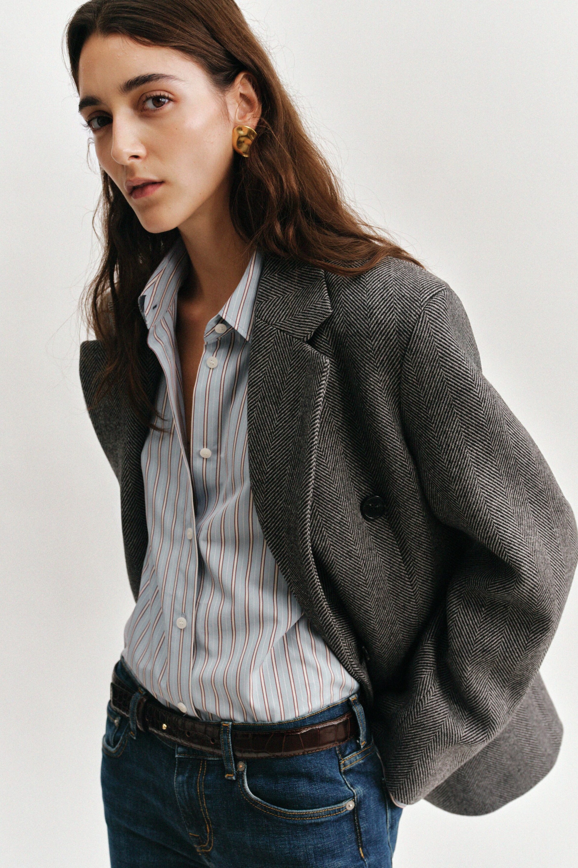Agrandir l'image: Un mannequin porte un blazer décontracté à double boutonnage en tissage chevron gris, avec revers crantés, présenté de trois quarts face. Le blazer est superposé à une chemise boutonnée en popeline de coton rayée bleu clair et rouge, rentrée dans un jean bleu. Une ceinture en cuir marron foncé brillant à motif croco, avec fermeture à rivet ton or, souligne la taille.