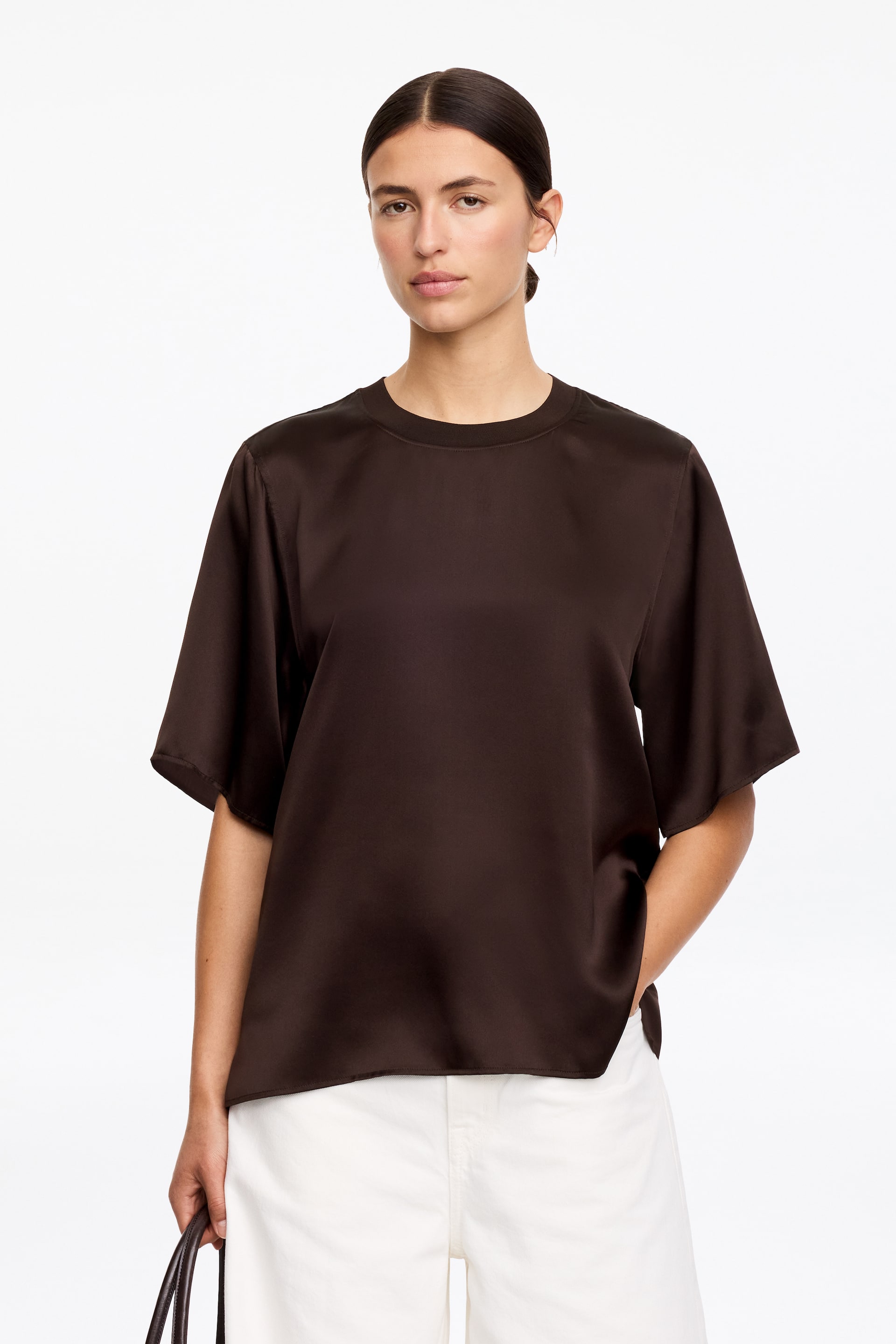 Visa större bild: En mörkbrun T-shirt i silkessatin, med ett flytande fall, subtil glans, ribbad rund hals och nedhasade axlar, bärs av modellen. Modellens högra hand vilar i en slits i sidosömmen.