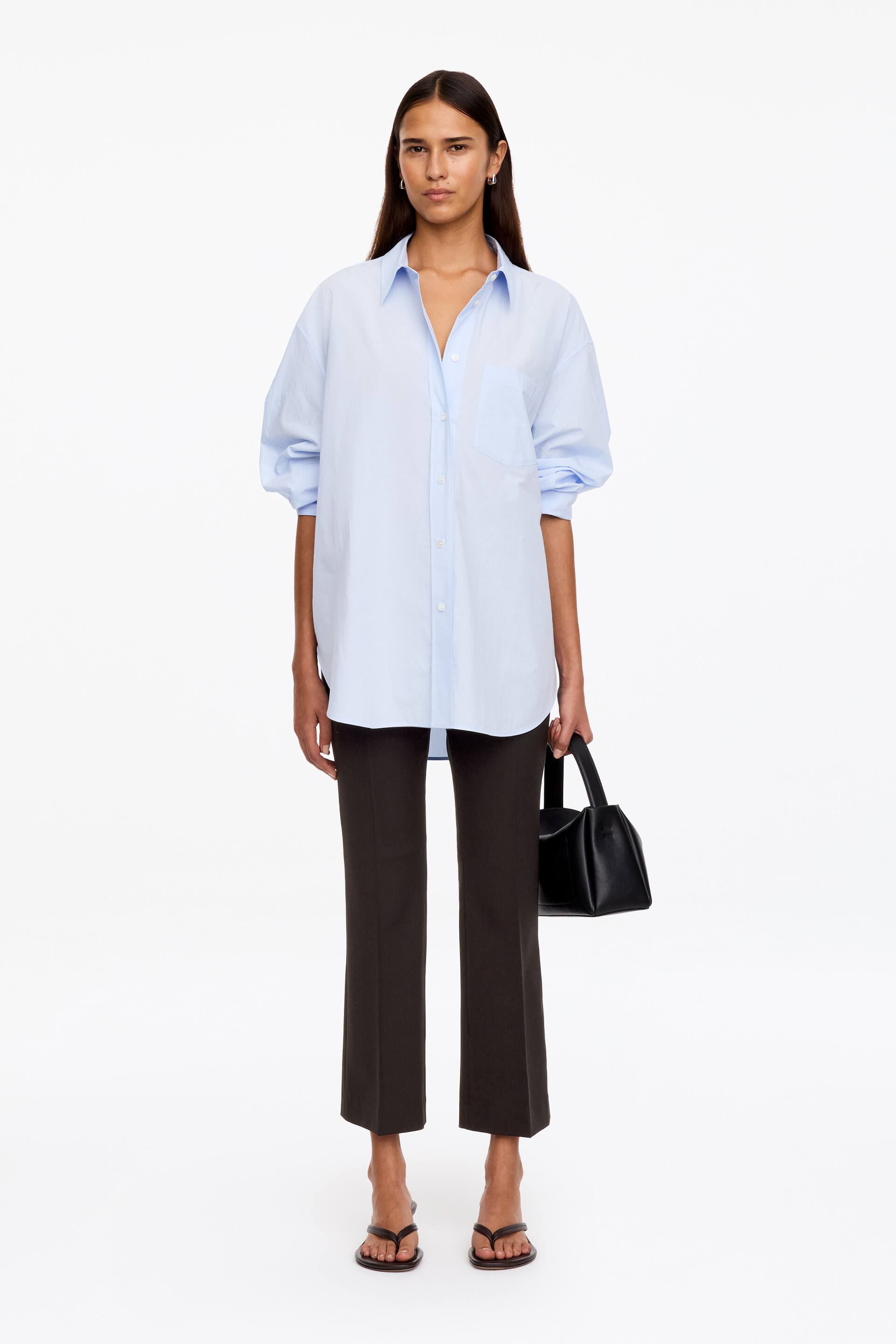 Ver imagen más grande: Estos pantalones de vestir ajustados de color marrón oscuro, con marcadas rayas de planchado delanteras y bajo al tobillo, los lleva la modelo. Los pantalones se combinan con una camisa oversize de botones en azul claro, llevada por fuera, y la modelo sostiene un pequeño bolso de mano negro.