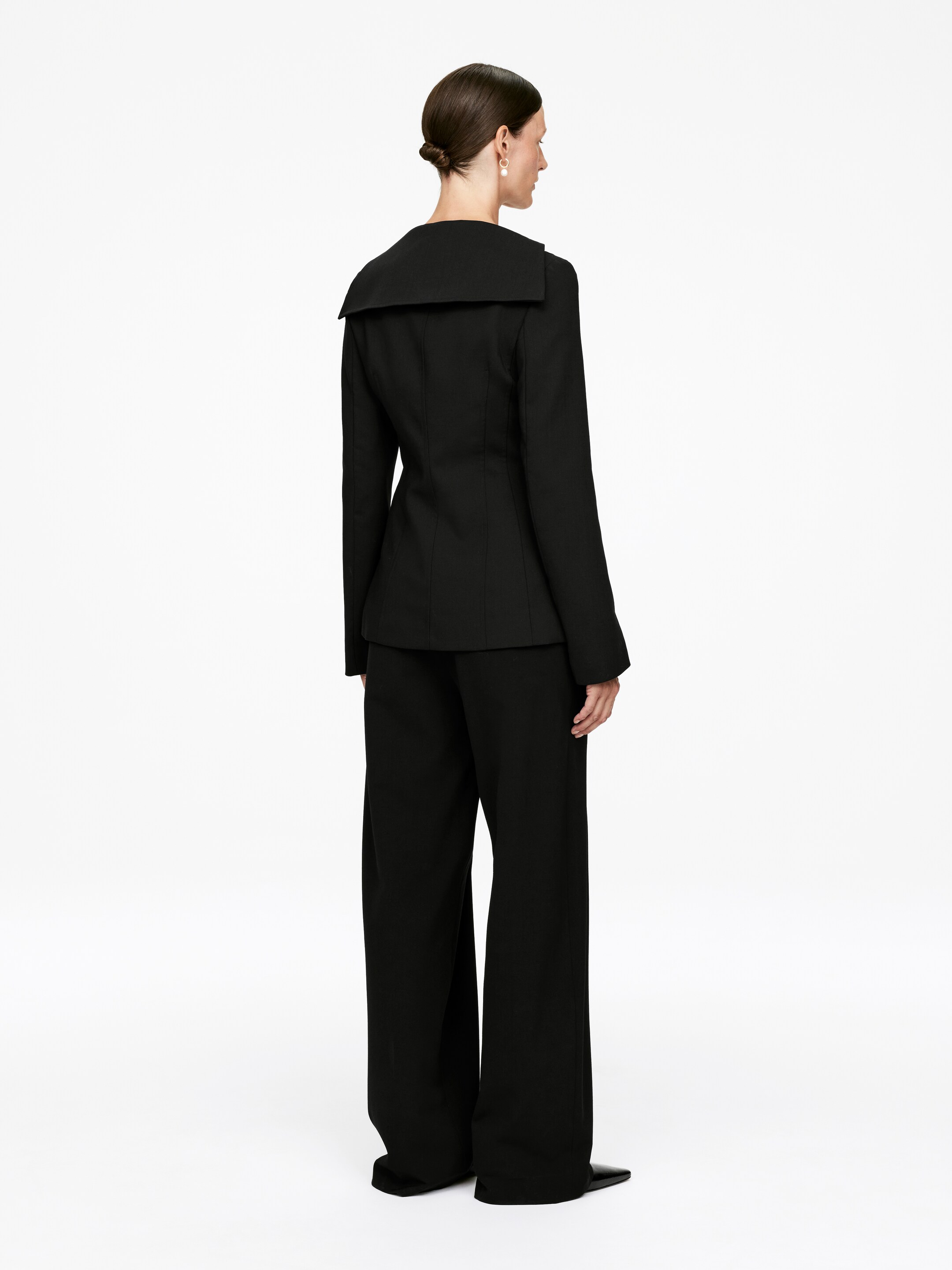 Visualizza immagine più grande: Un tailleur pantalone nero è esposto da una vista posteriore a tre quarti, mettendo in mostra la sua silhouette aderente e il design a gamba larga. La giacca presenta un ampio colletto strutturato che si estende sulle spalle, e i pantaloni appaiono in un taglio a lunghezza intera.