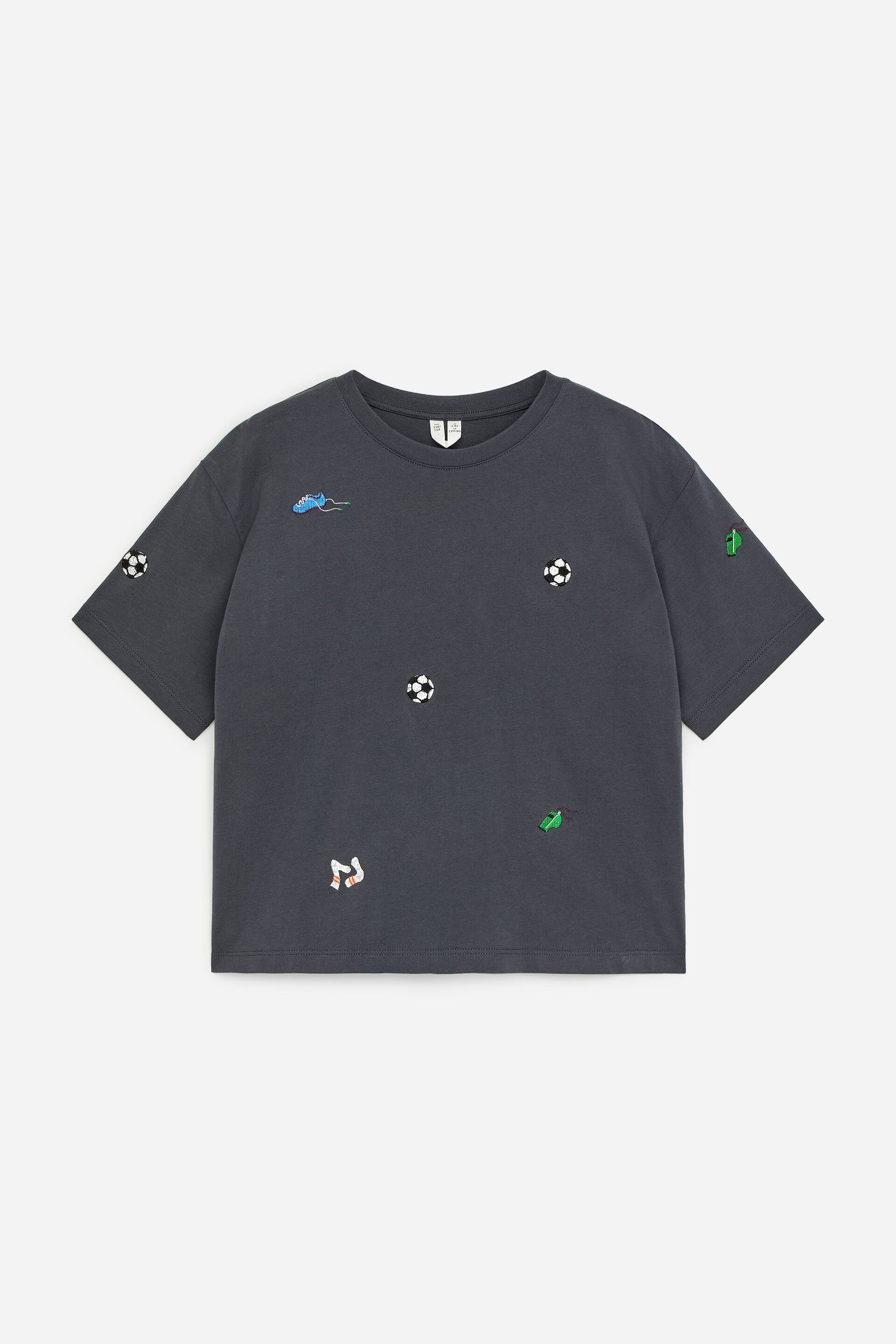 Größeres Bild anzeigen: Ein T-Shirt mit lockerer Passform, gefertigt aus weicher Baumwolle, wird von vorne präsentiert und zeigt eine dunkelgraue Farbe sowie einen gerippten Rundhalsausschnitt. Es ist mit einem Allover-Print versehen, der verstreute Fußbälle, grüne Fahnen, blaue Stollenschuhe und weiße sowie orangefarbene Socken zeigt.