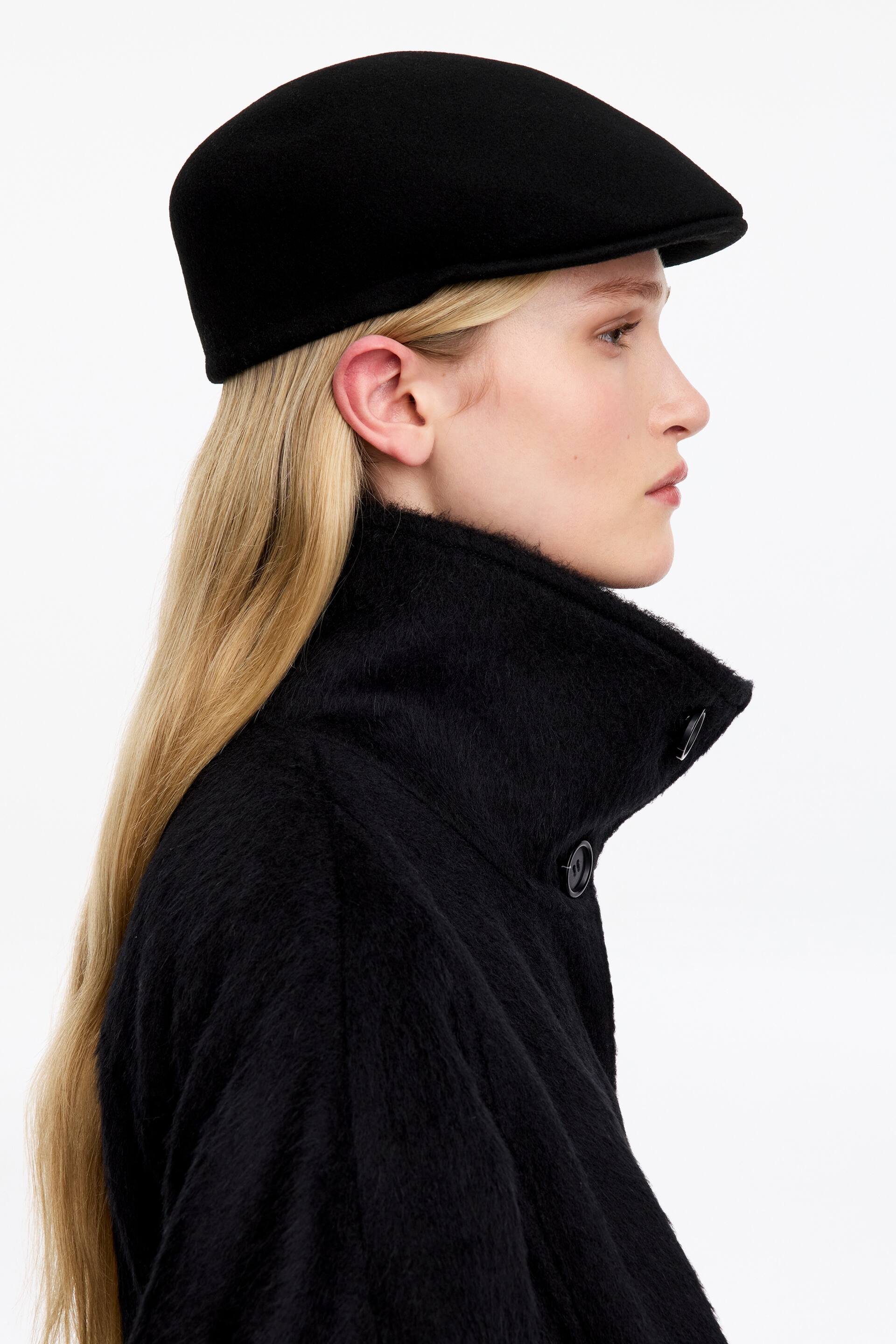 View larger image: A black wool flat cap with a structured, classic silhouette is being worn by the model, who is shown in profile. The cap is styled with a high-collared, textured black coat.