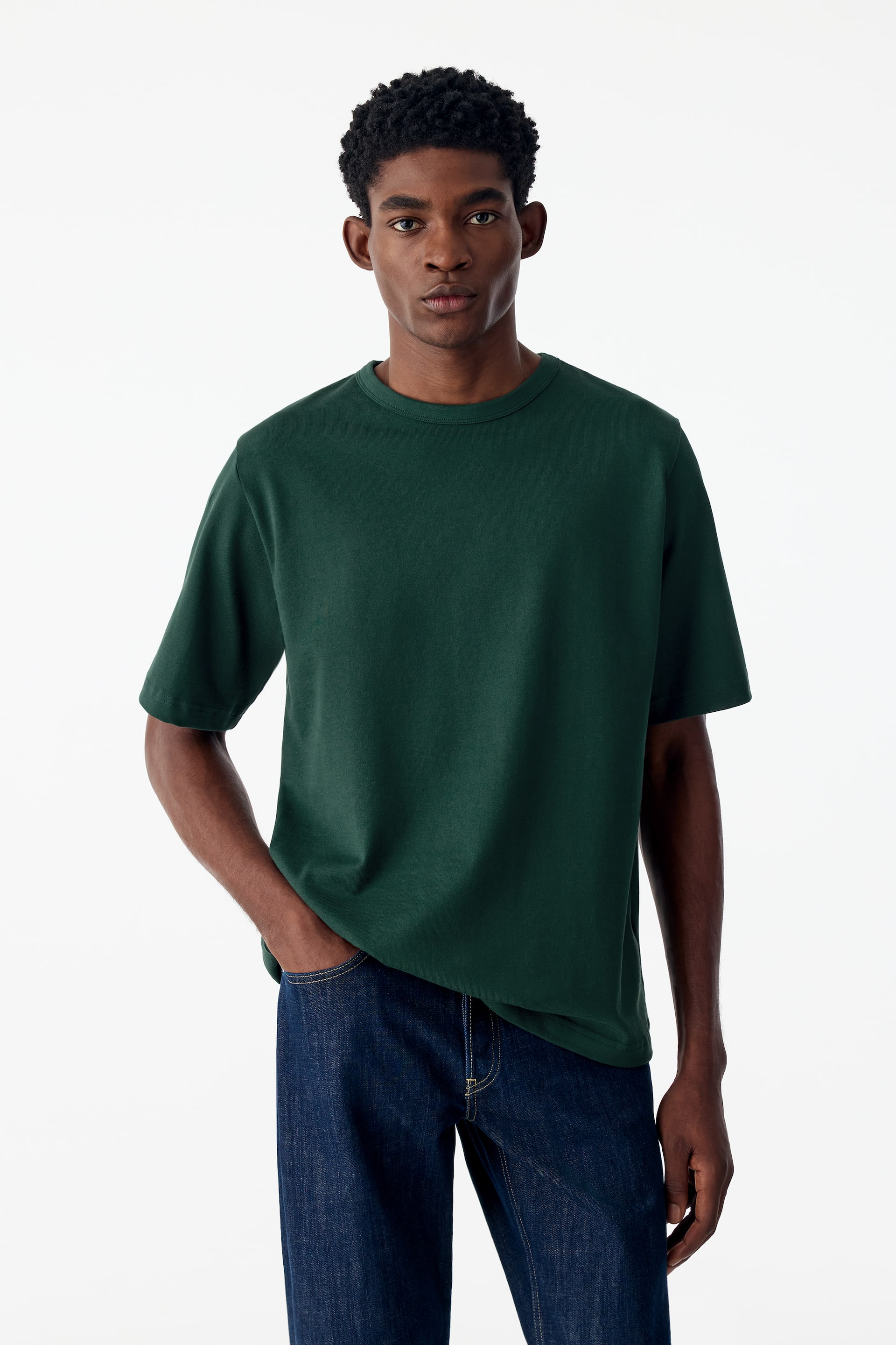 Se større billede: En mørkegrøn T-shirt med rund hals og kraftig kvalitet bæres af modellen og har en afslappet pasform, en ribstrikket halsudskæring og lidt længere korte ærmer. Modellen står med den ene hånd hvilende i lommen på mørkeblå jeans.
