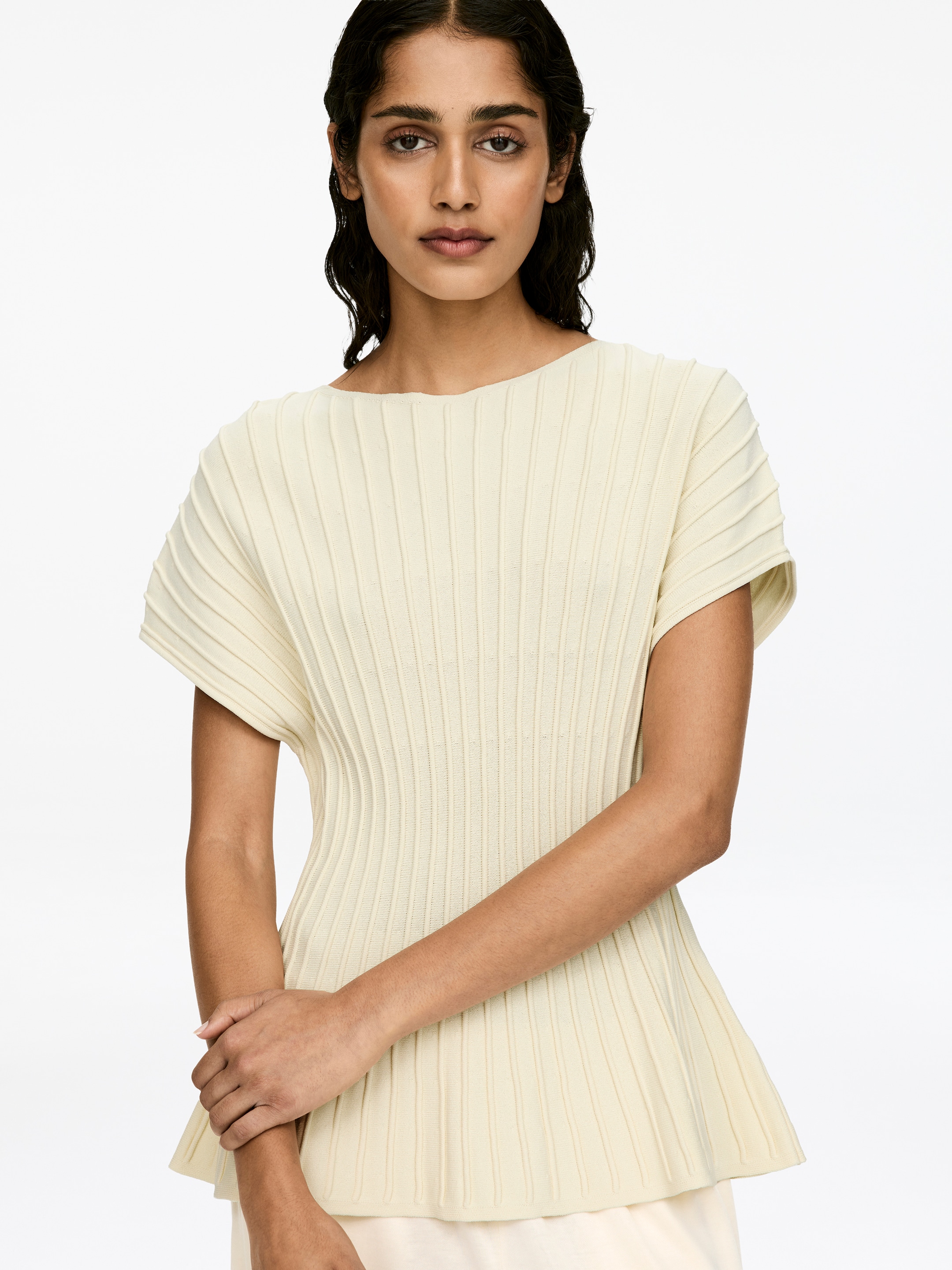 Visualizza immagine più grande: Un top a maglia fine color crema, caratterizzato da un punto ottomano orizzontale che crea una texture a coste verticali e una silhouette peplo, è indossato dalla modella. La modella incrocia le braccia davanti, con i pantaloni di colore chiaro visibili sotto l'orlo del top.