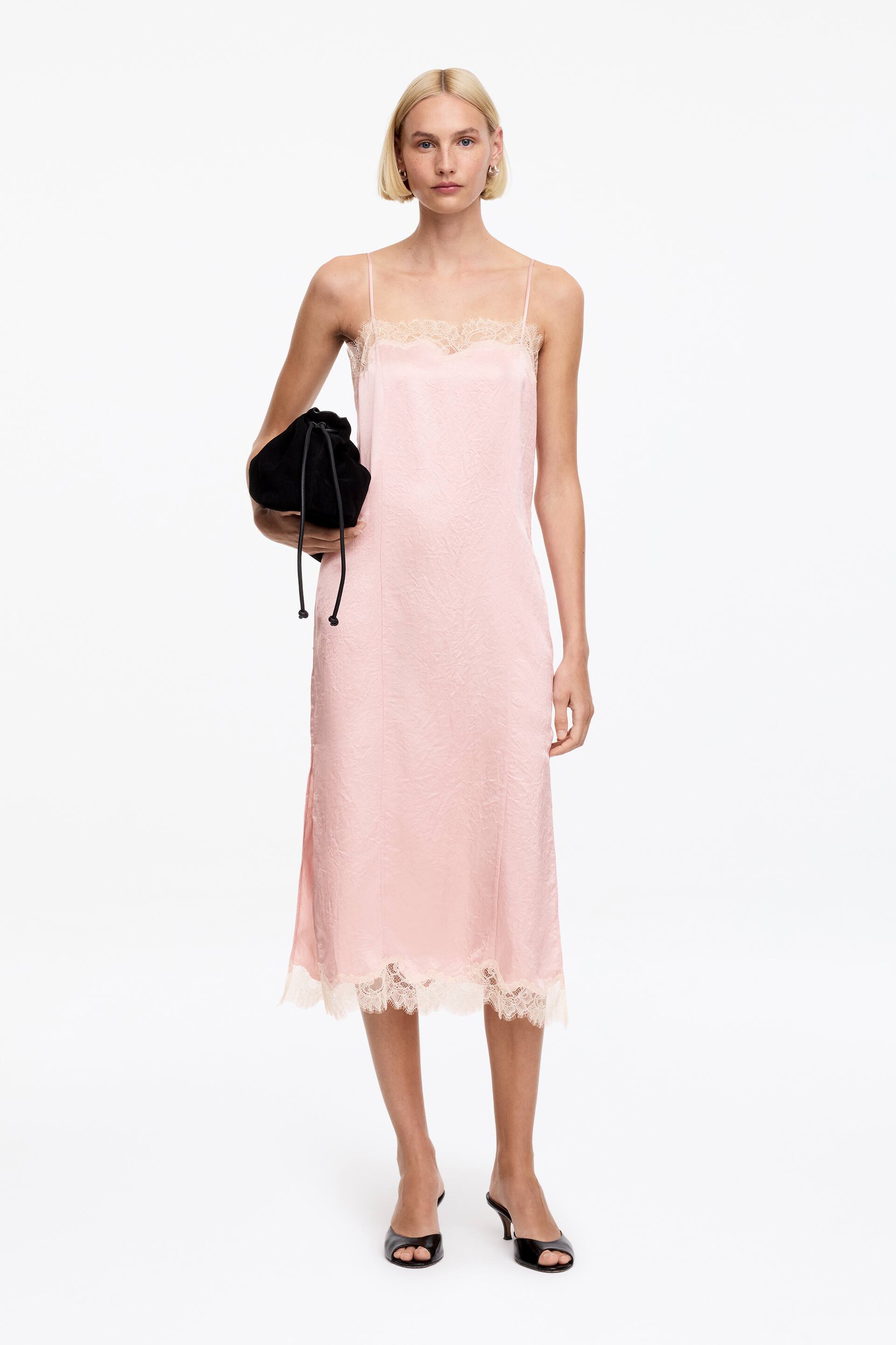 View larger image: The model is wearing a pink satin slip dress featuring a subtle textured pattern, delicate lace trim at the v-neckline and hem, and a mid-length regular fit. She is accessorised with a black drawstring clutch bag and black open-toe mules.