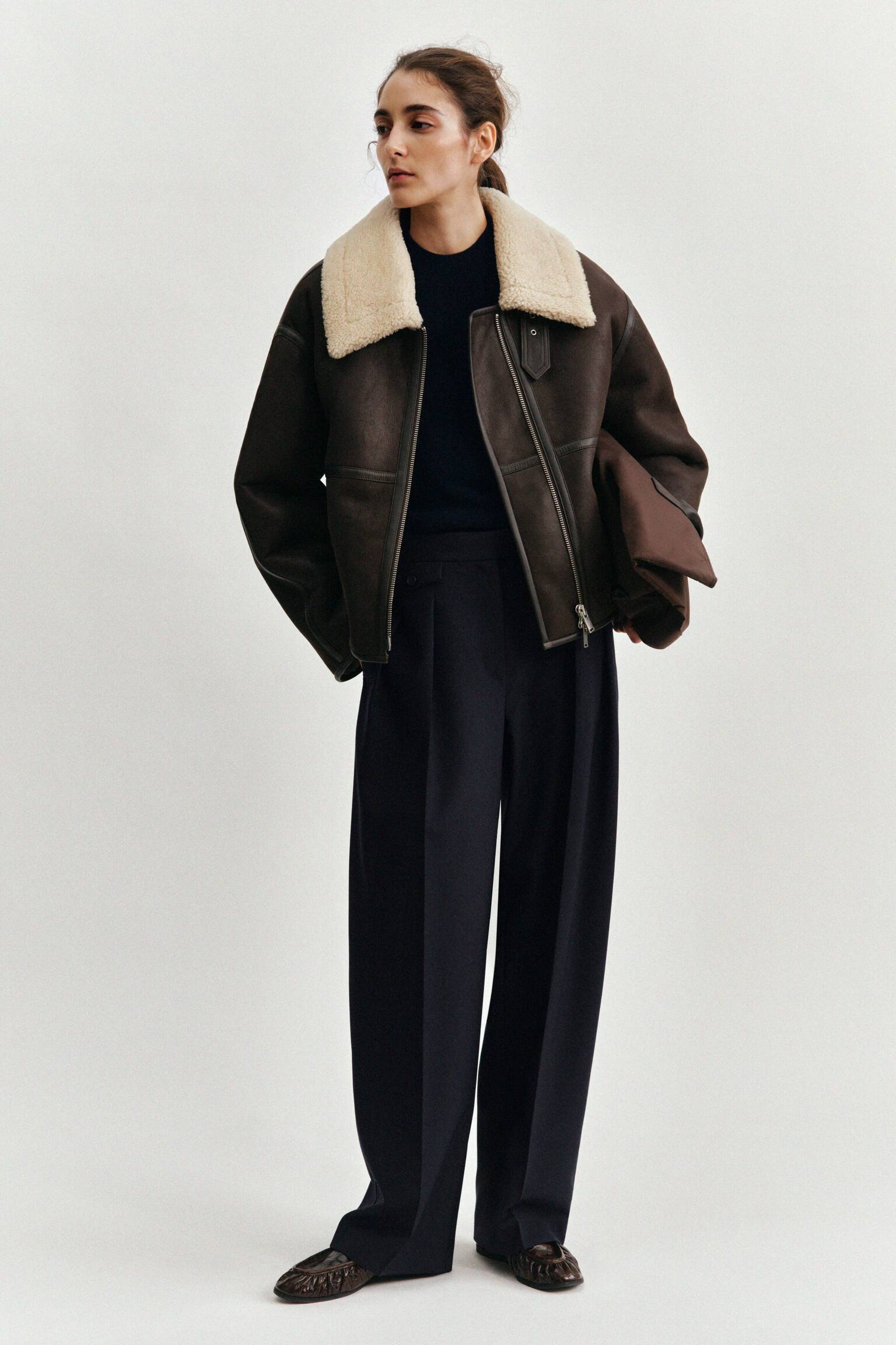 En modell visar en mÃ¶rk marinblÃ¥ stickad jumper med mock-neck, lager under en brun jacka med shearling-krage, kombinerad med hÃ¶ga, vida marinblÃ¥ skrÃ¤ddarsydda byxor med skarpa pressveck framtill. Looken kompletteras med mÃ¶rkbruna loafers i croco-prÃ¤glat lÃ¤der med rynkade detaljer och en brun tote bag i nylon som hÃ¥lls under armen. - 1