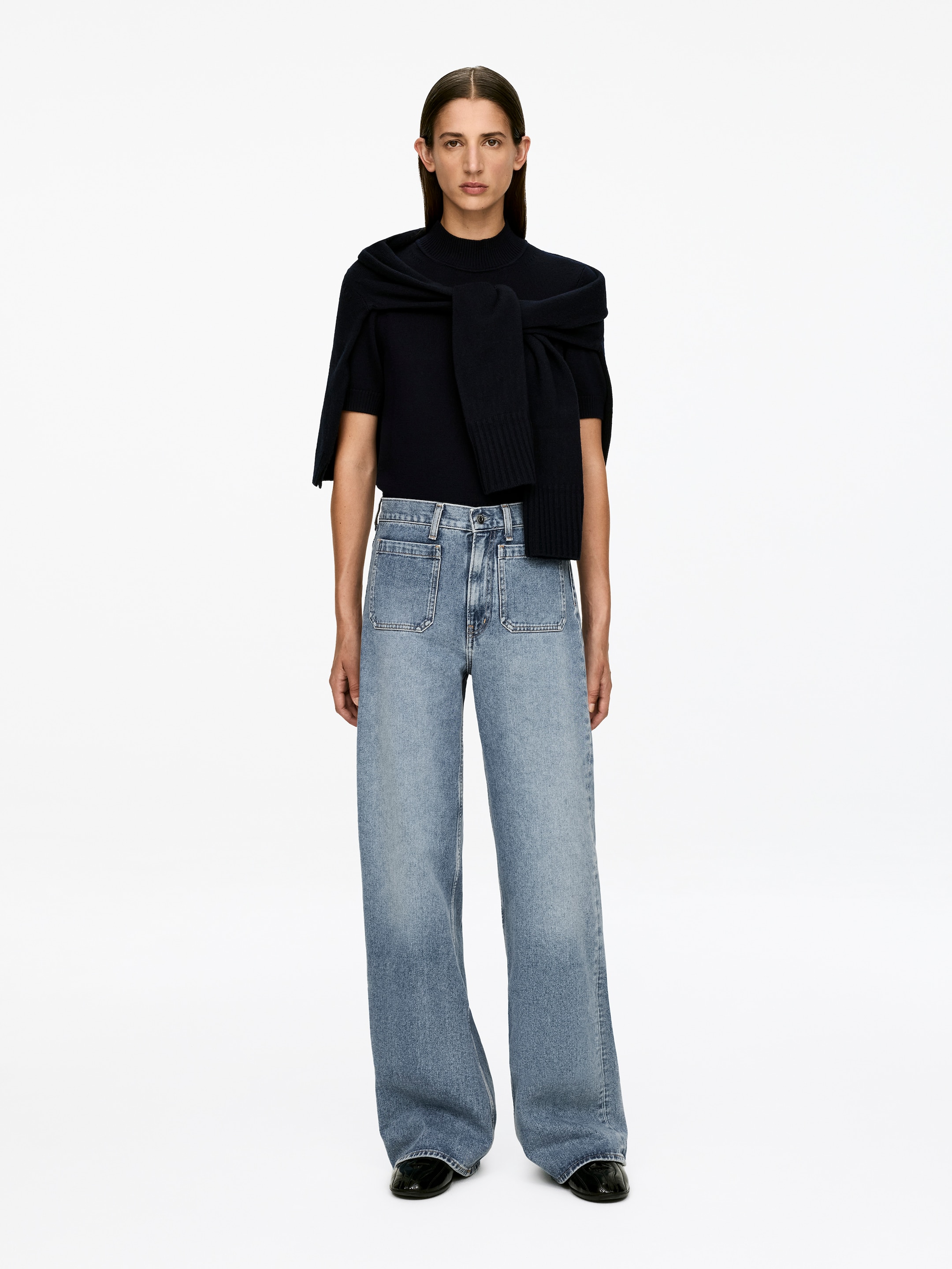 Visualizza immagine più grande: Il denim LUPINE azzurro chiaro è indossato dalla modella, con una vita alta, una vestibilità aderente attraverso la coscia e gambe svasate, con visibili tasche frontali applicate e una chiusura con zip. La modella sta abbinando il denim con un top scuro a maniche corte e un maglione scuro drappeggiato sulle spalle.