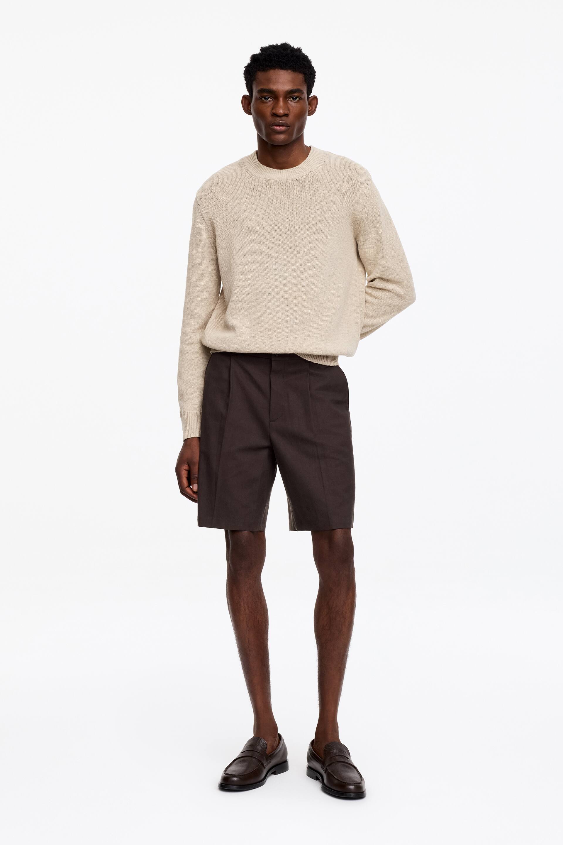 Grotere afbeelding bekijken: Een licht beige trui met ronde hals en een subtiele breistructuur, geribbelde boorden en zoom, valt losjes over donkerbruine geplooide shorts die net boven de knie komen.