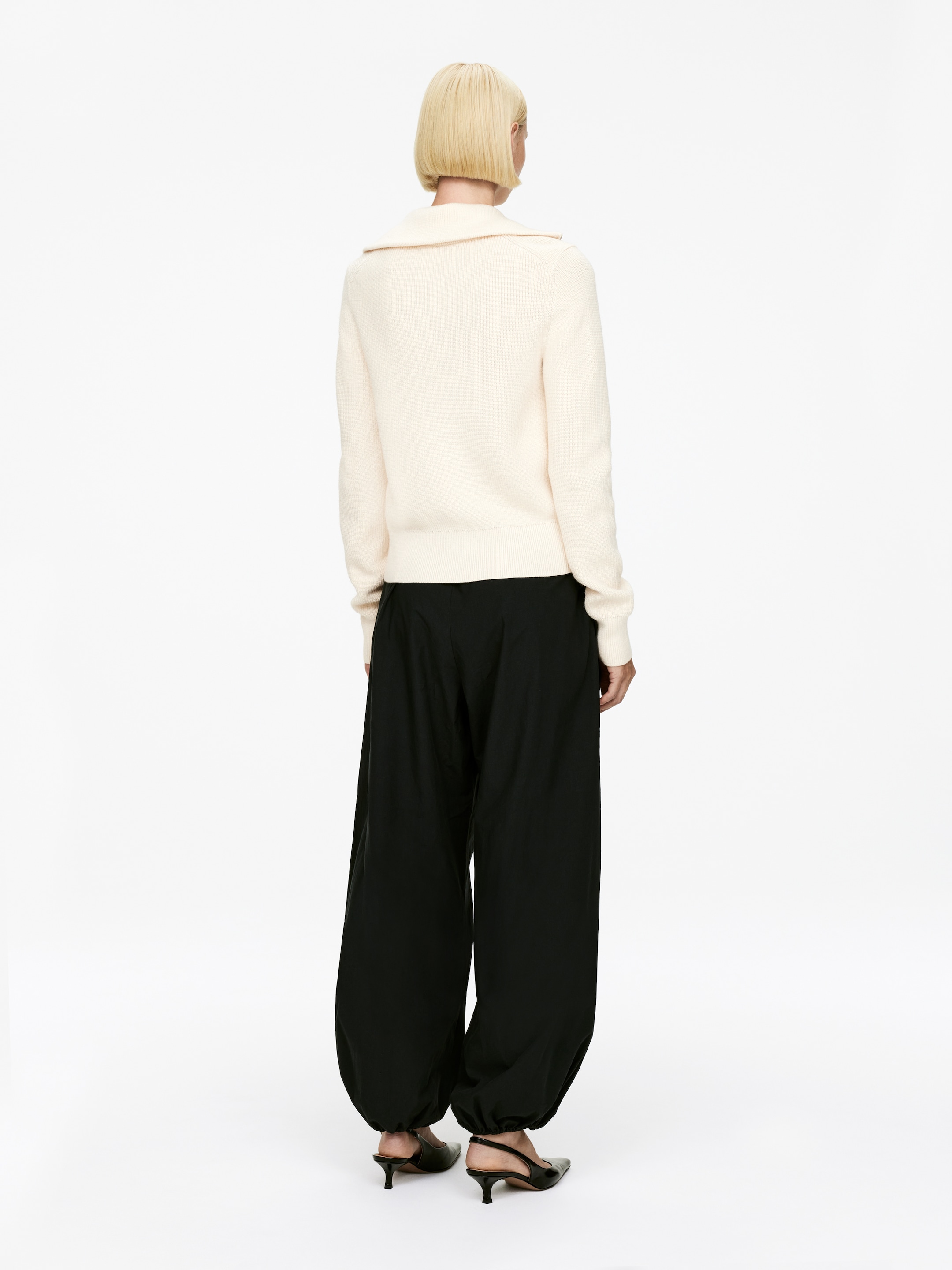 Agrandir l'image: Un pull tricoté de couleur crème est présenté de dos, avec un large col évasé côtelé et un ourlet côtelé. Il est porté avec un pantalon noir à jambes larges qui se rassemble aux chevilles, créant une silhouette en ballon, et des escarpins slingback noirs à petits talons.