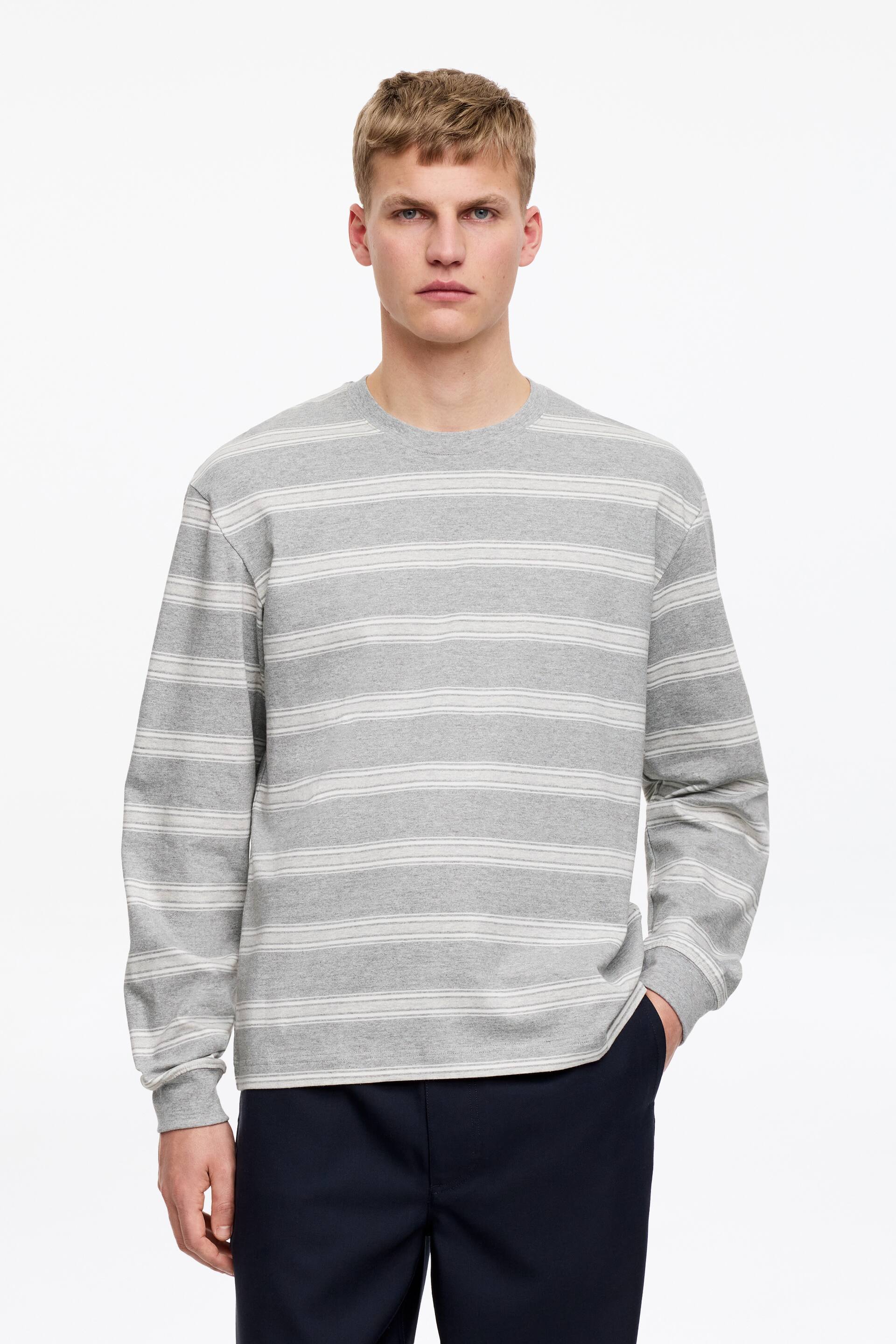 Grotere afbeelding bekijken: Een grijs T-shirt met lange mouwen en horizontale witte strepen, met geribde boorden aan de ronde hals en de mouwen, wordt door het model in een relaxte pasvorm gedragen. Het model draagt bovendien een donkere broek.