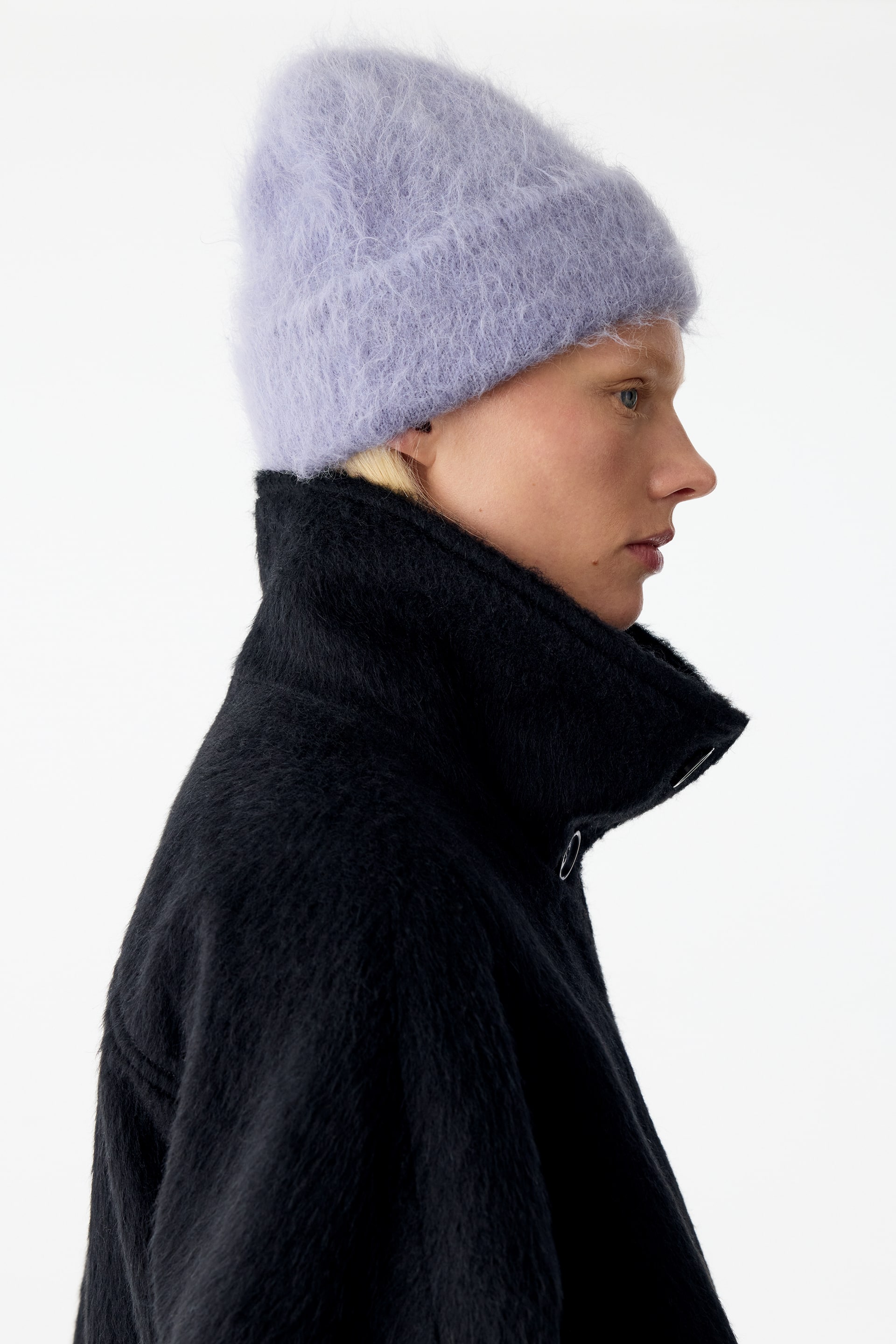 View larger image: A fluffy, lilac knitted beanie with a folded cuff is being worn by the model, who is facing to the side. The beanie is styled with a high-collared, textured black coat.