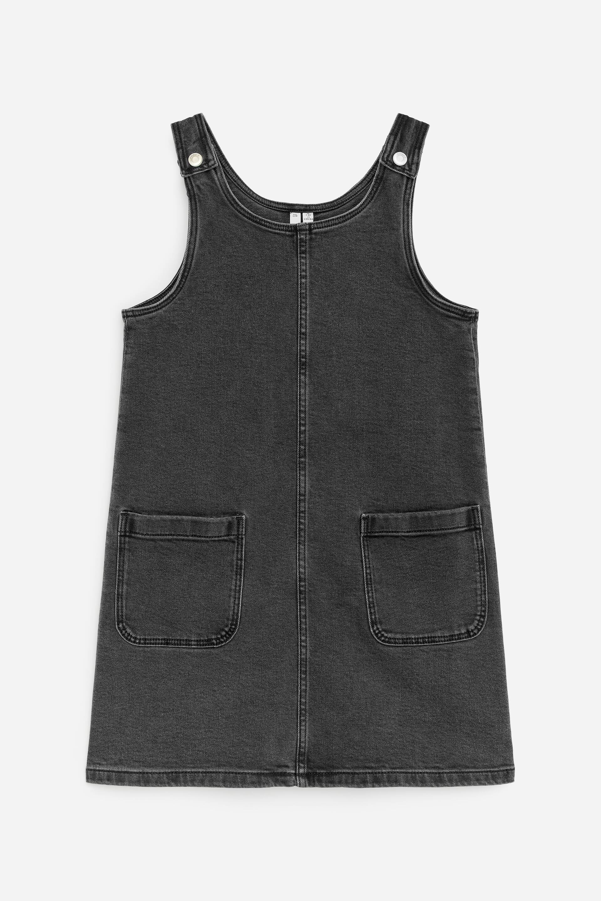 Visa större bild: En mörkgrå denimförklädesklänning visas FRAM, med en rundad halsringning, två axelband som är fästa med silverfärgade knappfästen, en central vertikal söm och två stora påsydda fickor placerade nära fållen.