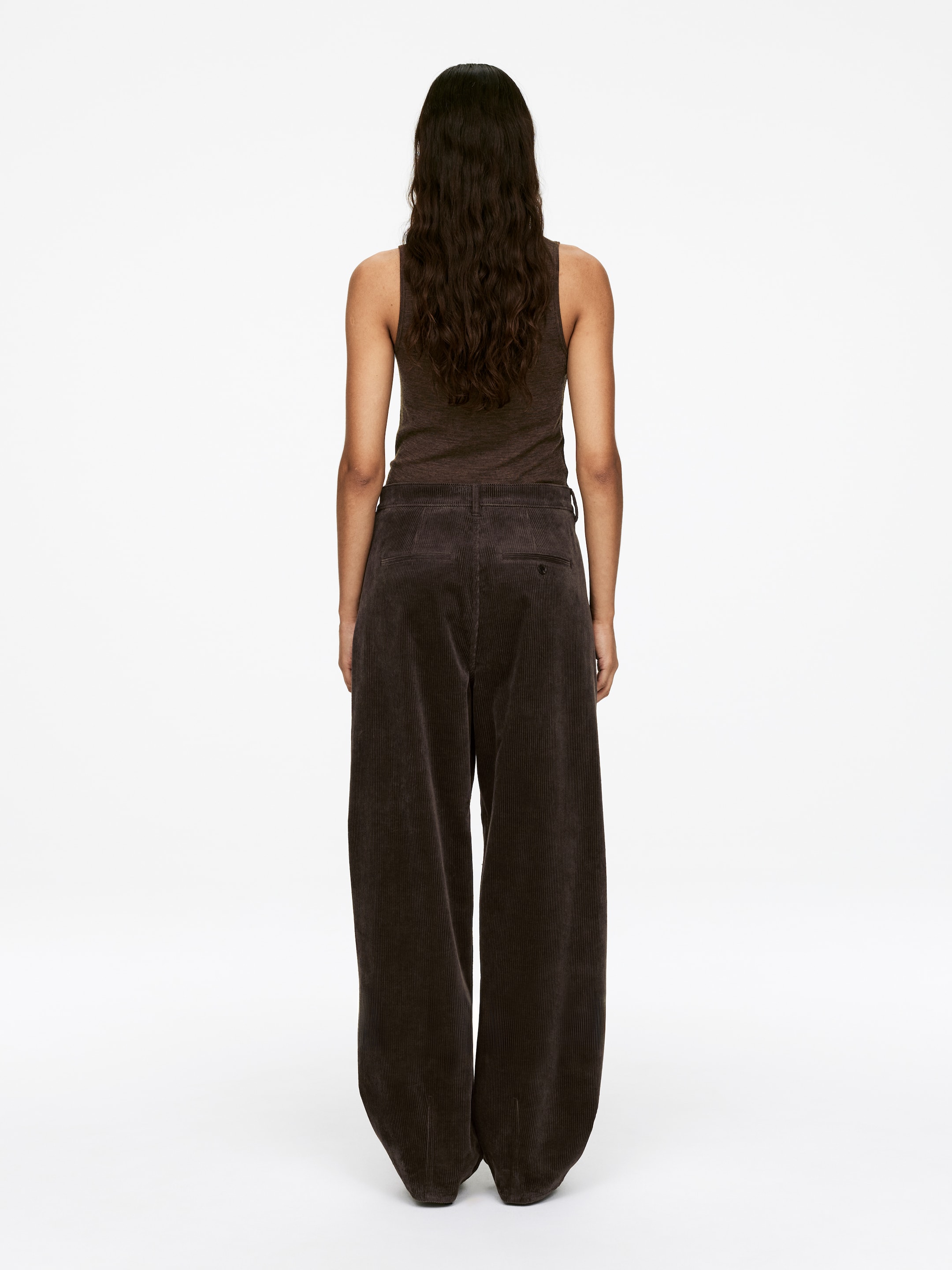 Agrandir l'image: Le mannequin porte un pantalon en velours côtelé marron foncé à jambes larges, avec une taille mi-haute et une seule poche passepoilée à l'arrière.