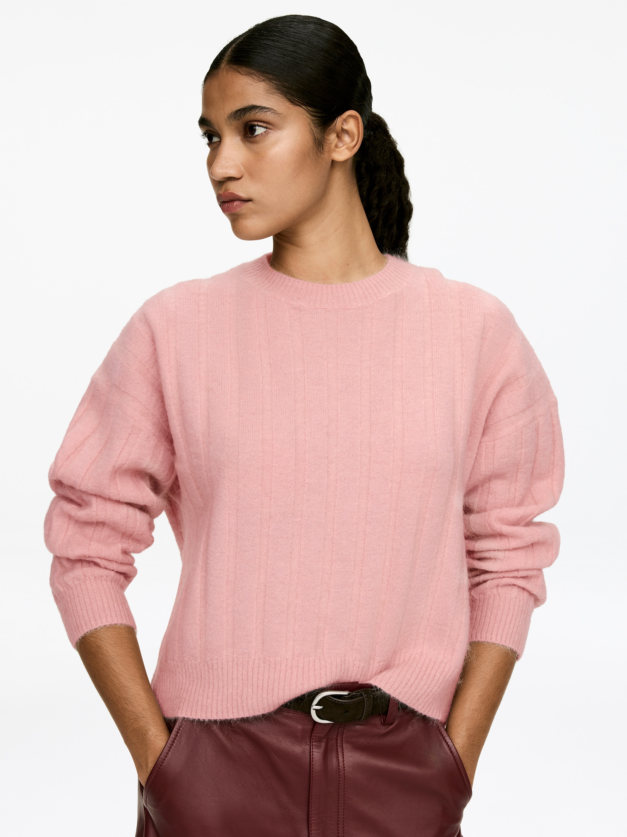 Grösseres Bild ansehen: Ein pinker Pullover mit breiter Rippenstruktur und flauschiger Textur, ausgestattet mit Rundhalsausschnitt und langen Ärmeln, wird vom Model getragen. Der locker sitzende Pullover ist über bordeauxroten Hosen gestylt, wobei die Hände des Models in den Taschen ruhen und ein dunkler Gürtel sichtbar ist.