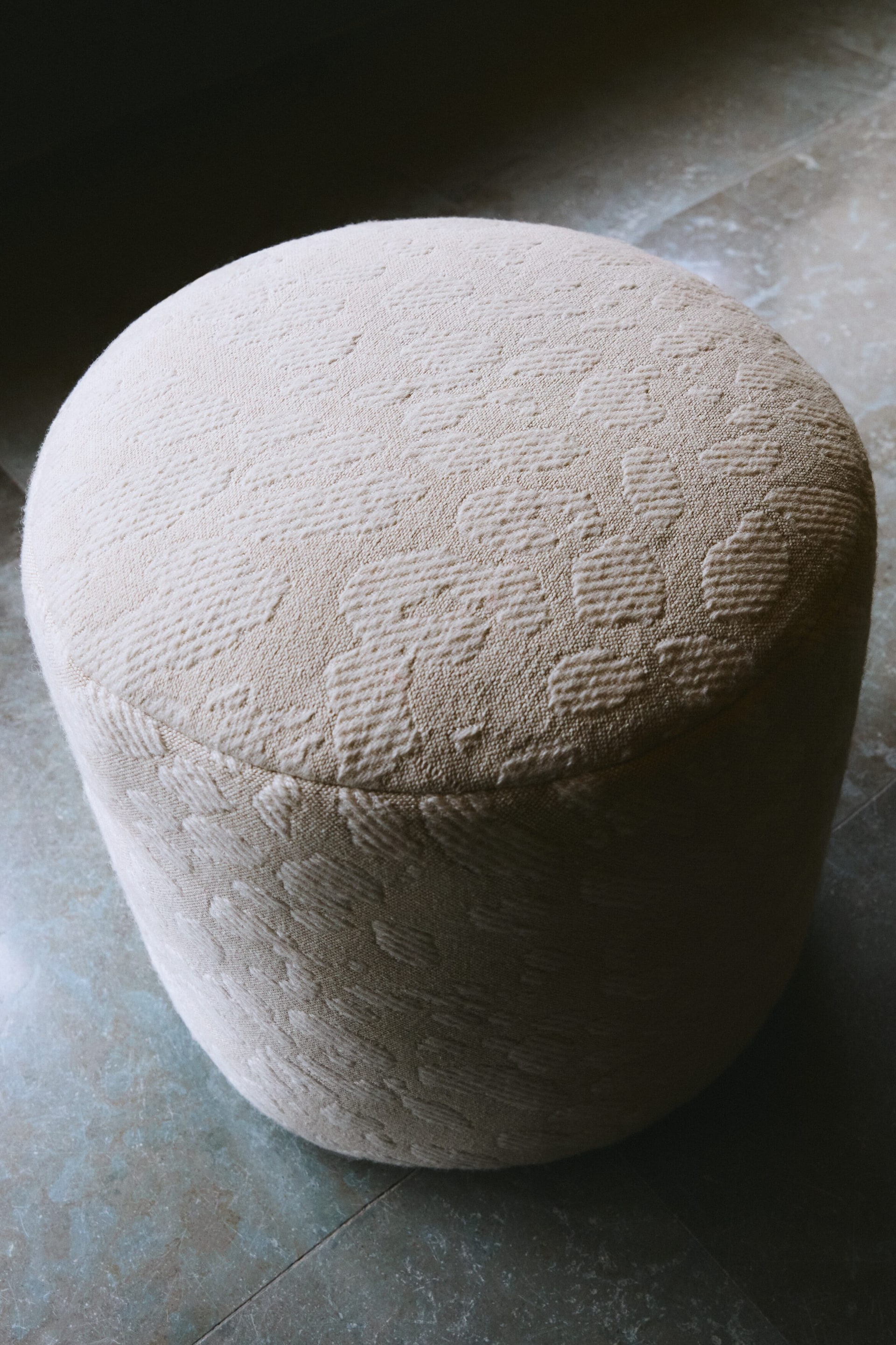View larger image: A light cream cylindrical ottoman is being presented from a slightly elevated, downward angle. It features a textured, woven fabric with a raised, repeating abstract pattern covering its gently rounded top surface and sides.
