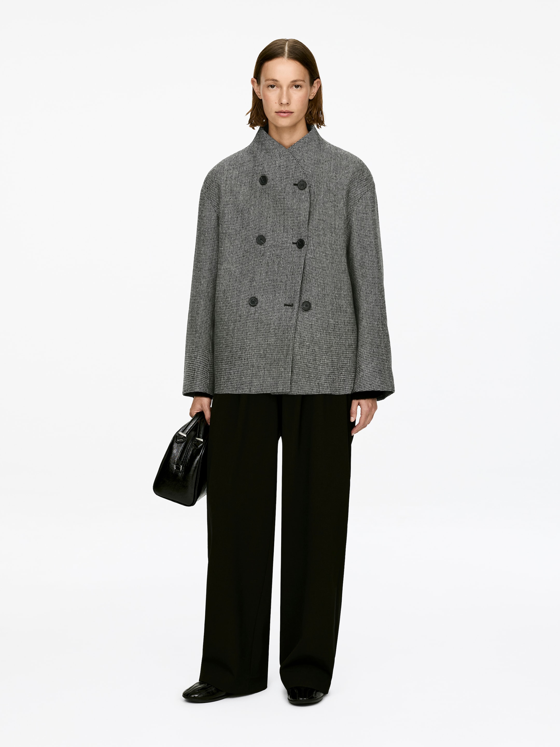 Grösseres Bild ansehen: Eine grau-weiß gemusterte zweireihige Jacke, gefertigt aus einer Wollmischung mit einer übergroßen Silhouette, hohem Stehkragen und schwarzen Knöpfen, wird vom Model getragen, das steht und eine schwarze Handtasche hält.