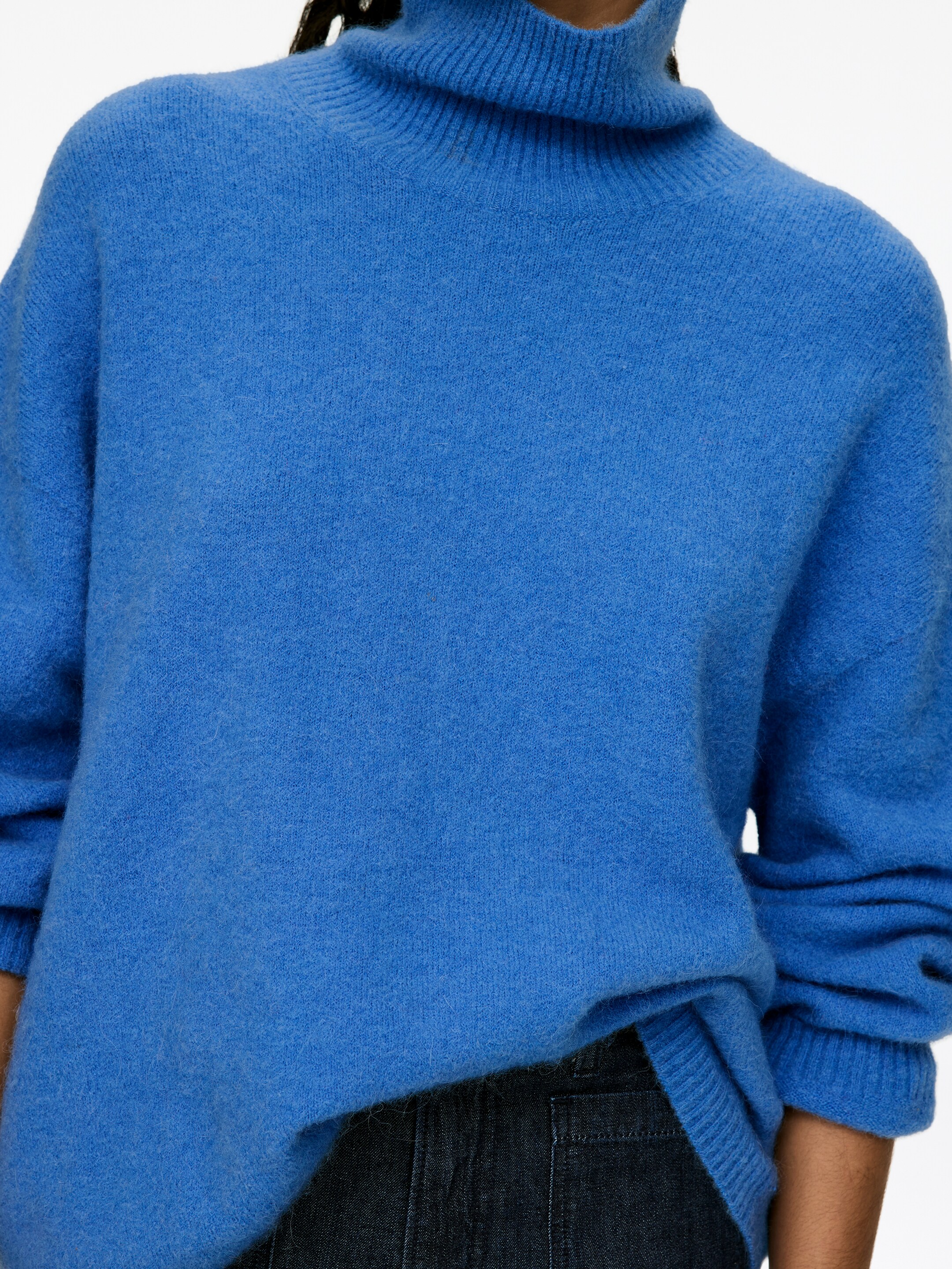 Vis større bilde: En blå turtleneck-genser blir presentert fra en front-side vinkel, med en ribbestrikket rullekrage, en myk, litt lodden strikket tekstur, og lange ermer som ender i ribbestrikkede mansjetter, der den avslappede kanten delvis henger over et mørkt plagg.