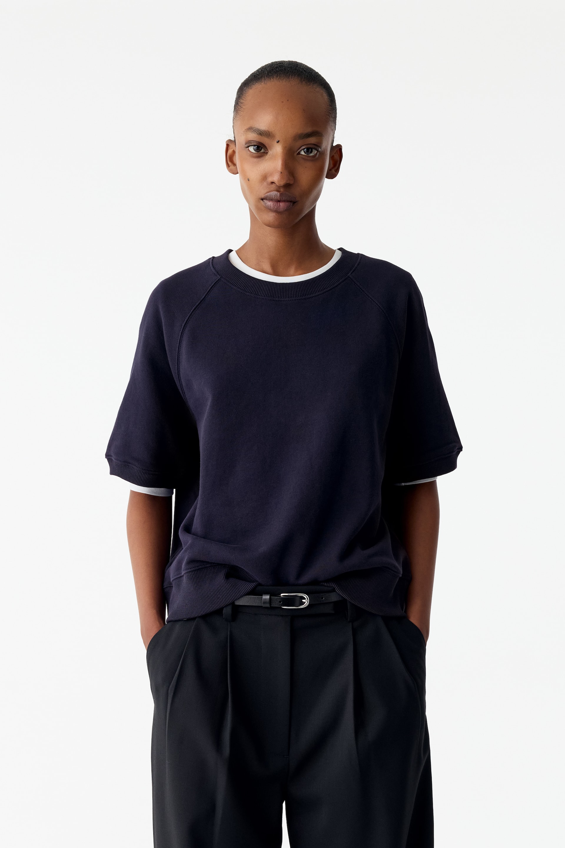 Agrandir l'image: Le mannequin porte un sweat-shirt à manches courtes bleu marine foncé, superposé sur un t-shirt blanc. Ce modèle à col rond présente des manches raglan et des finitions côtelées au niveau de l’encolure, des poignets et de l’ourlet.
