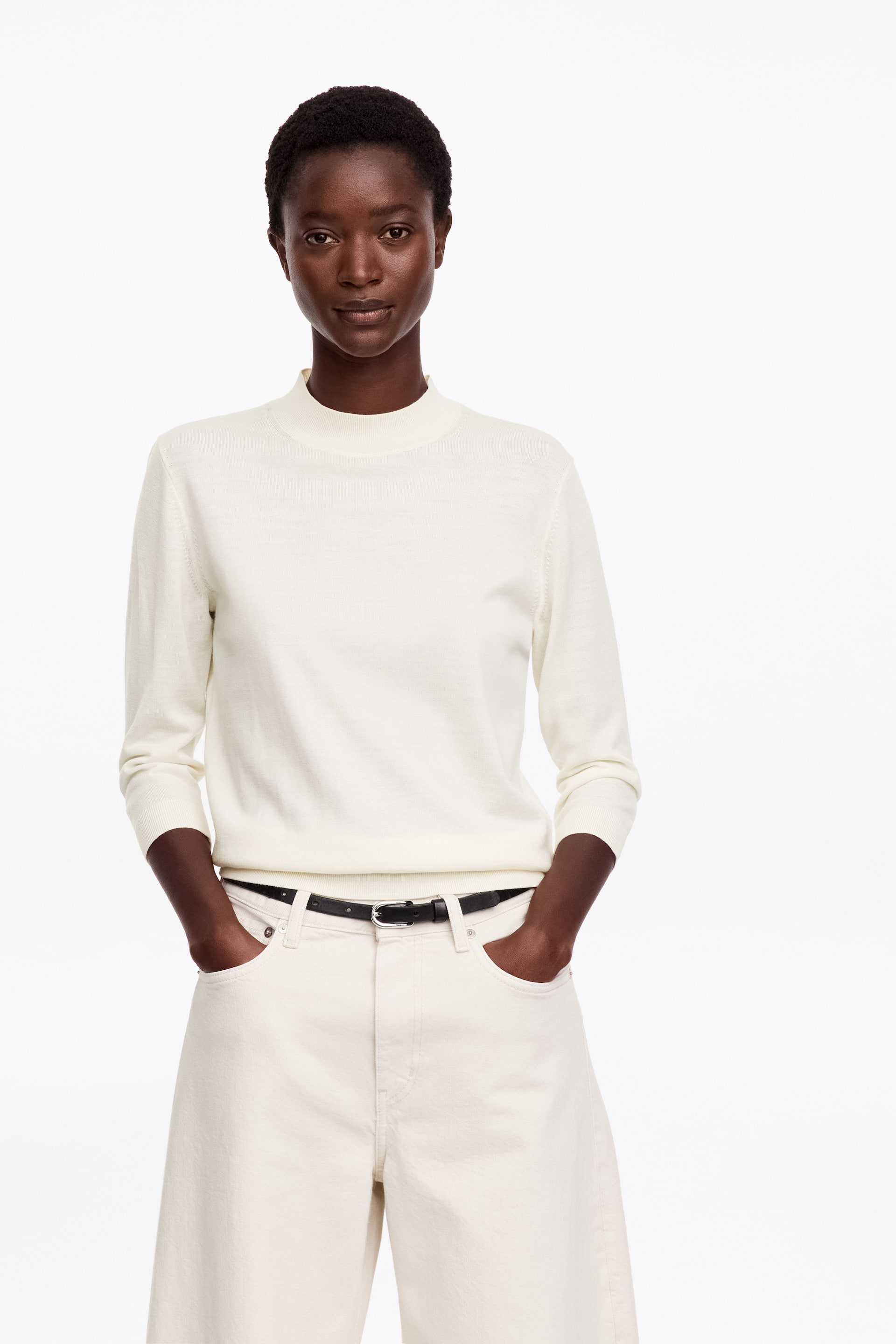 Grotere afbeelding bekijken: Een crèmekleurige top van merinowol met een opstaande, nepkraag en driekwartmouwen wordt gedragen door het model. De top is gestyled met bijpassende gebroken witte broek en een dunne zwarte riem.