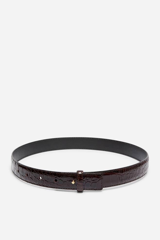 Une ceinture en cuir marron foncÃ©, dotÃ©e dâune texture brillante Ã  motif crocodile, est prÃ©sentÃ©e de face, disposÃ©e en boucle circulaire. La ceinture arbore une petite fermeture Ã  clou ton or, fixÃ©e dans lâun de ses trous de rÃ©glage et maintenue en place par un passant assorti en cuir. - 1
