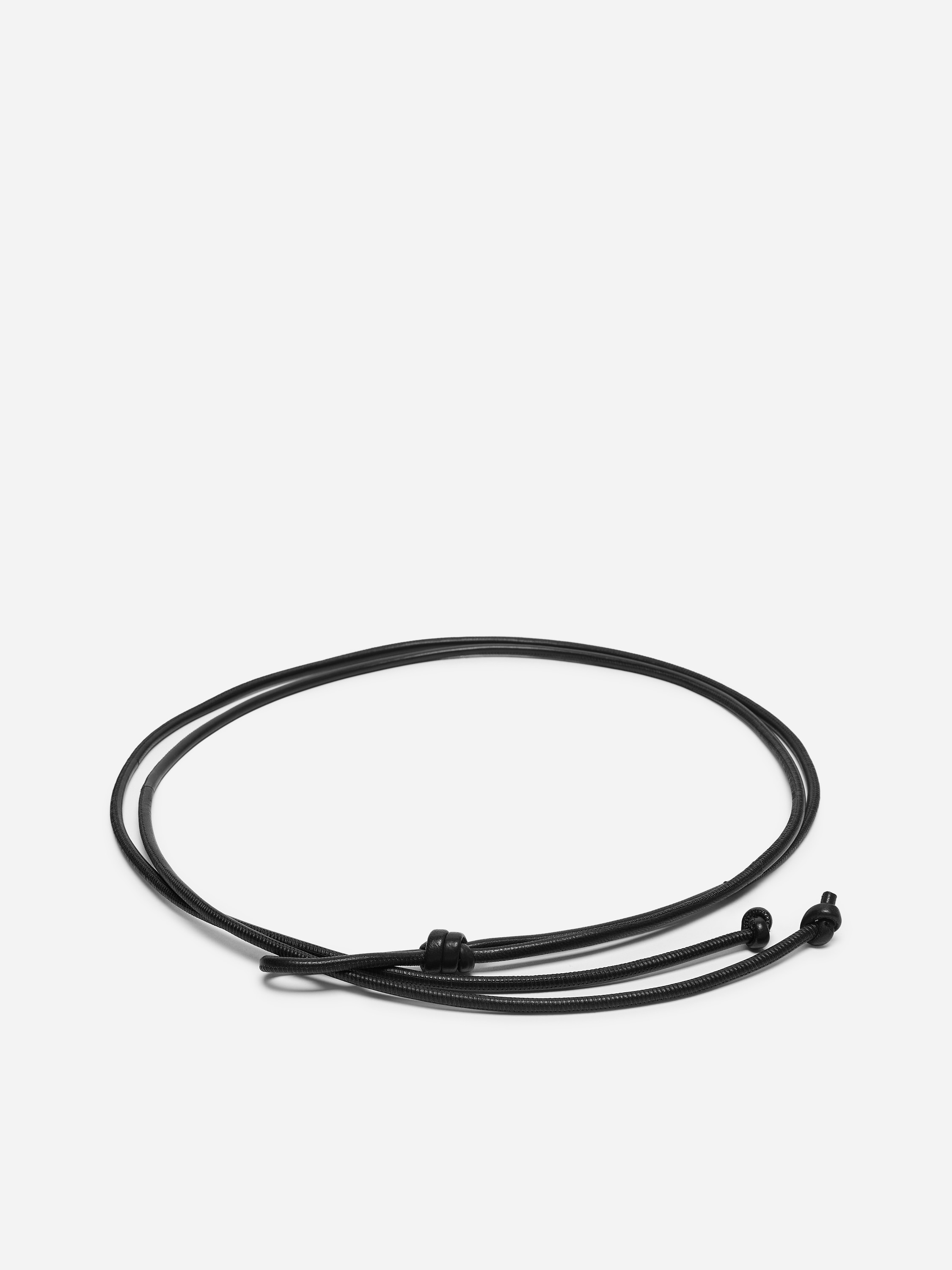 Una sottile cintura da vita in pelle nera con dettagli annodati e lunghe estremità sottili pendenti è indossata dalla modella sopra una tunica nera a maniche lunghe. La cintura è annodata al centro sul davanti, stringendo il capo in vita.
