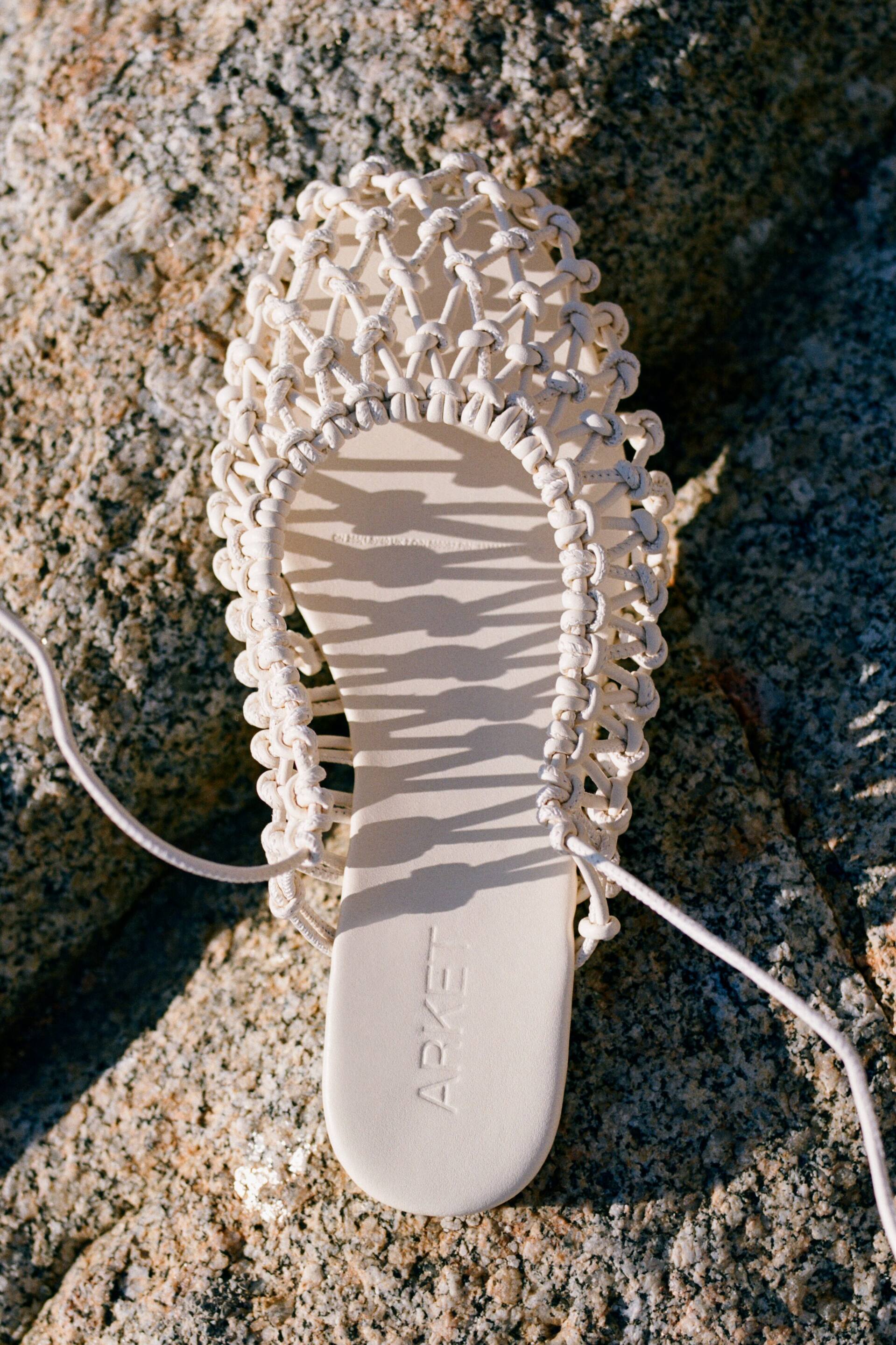 Grotere afbeelding bekijken: Een off-white platte sandaal met een ingewikkeld macramé-geknoopt bovendeel dat een open geweven ontwerp vormt, met lange dunne veters die aan de zijkanten tevoorschijn komen. De platte binnenzool toont de in reliëf gedrukte merknaam "ARKET" nabij de hiel.
