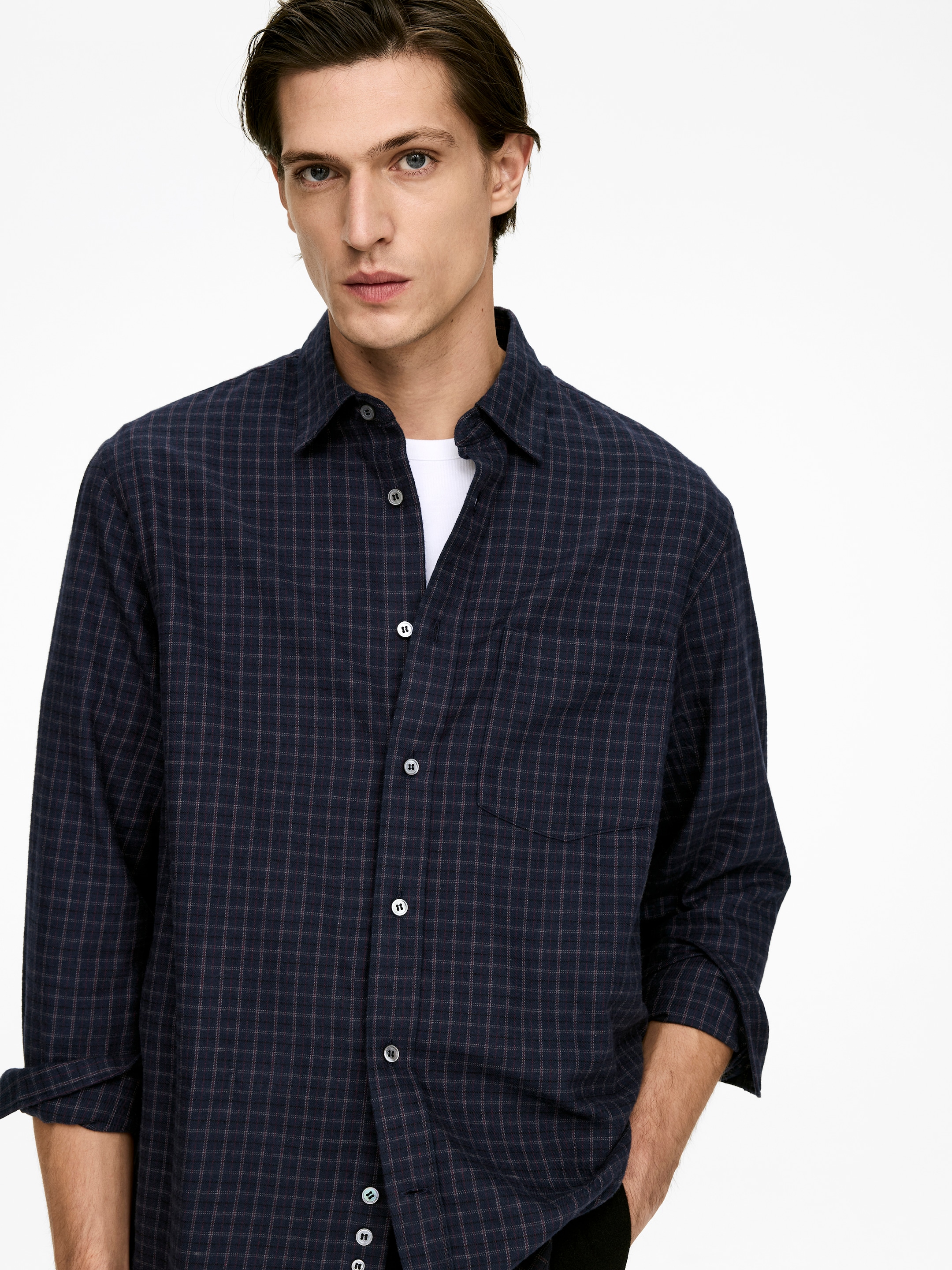 Visualizza immagine più grande: Una camicia a maniche lunghe di colore blu scuro viene presentata, con un motivo a griglia sottile, un colletto standard e una chiusura frontale con bottoni chiari. Le maniche sono arrotolate fino agli avambracci, una tasca applicata è visibile sul petto sinistro e sotto è indossata una t-shirt bianca.