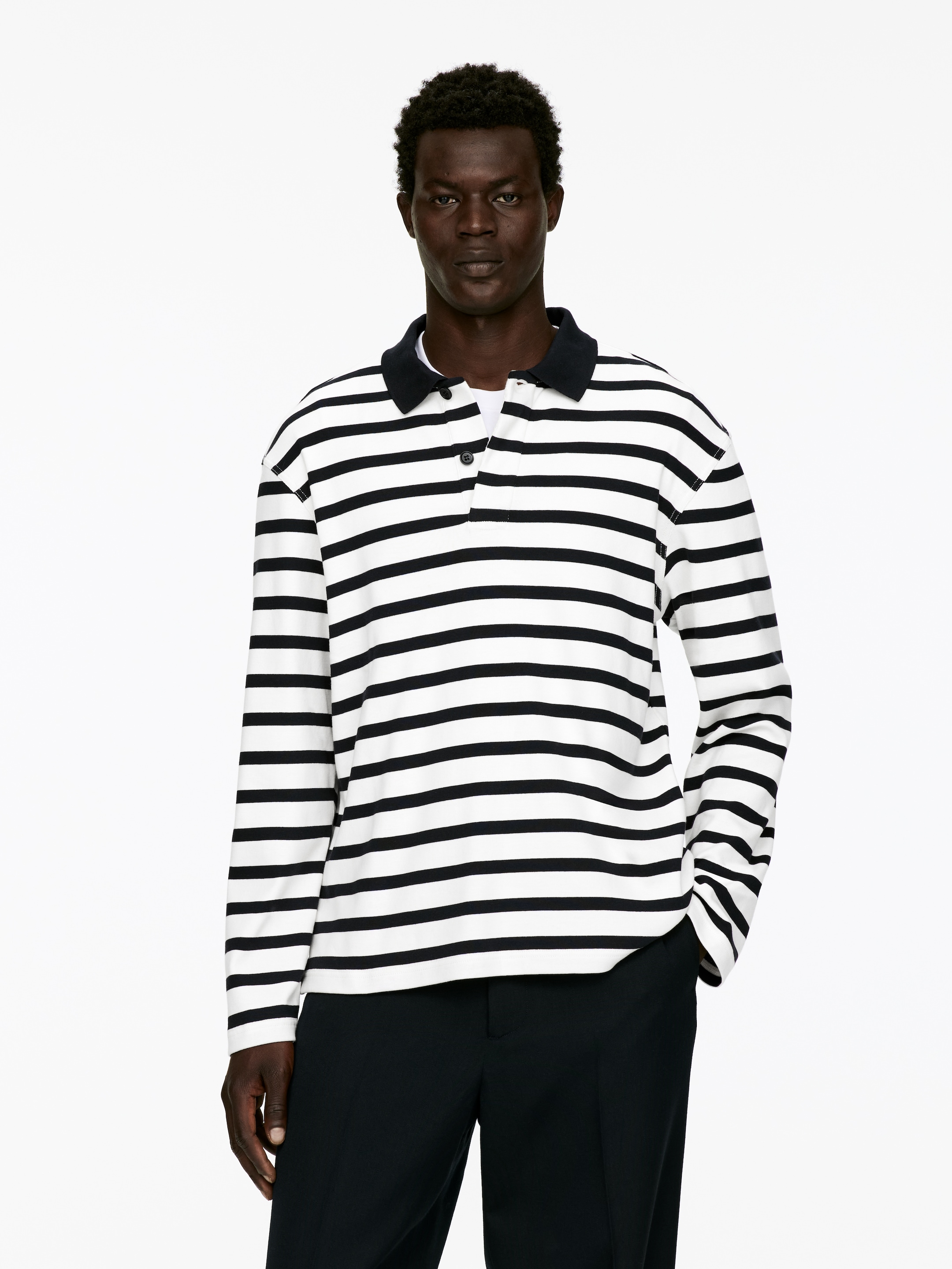 Se større billede: En langærmet poloshirt med sorte og hvide vandrette striber, en mørk ribbet krave og en halv placket med en synlig knap bæres af modellen. En hvid undertrøje er synlig under poloens placket.