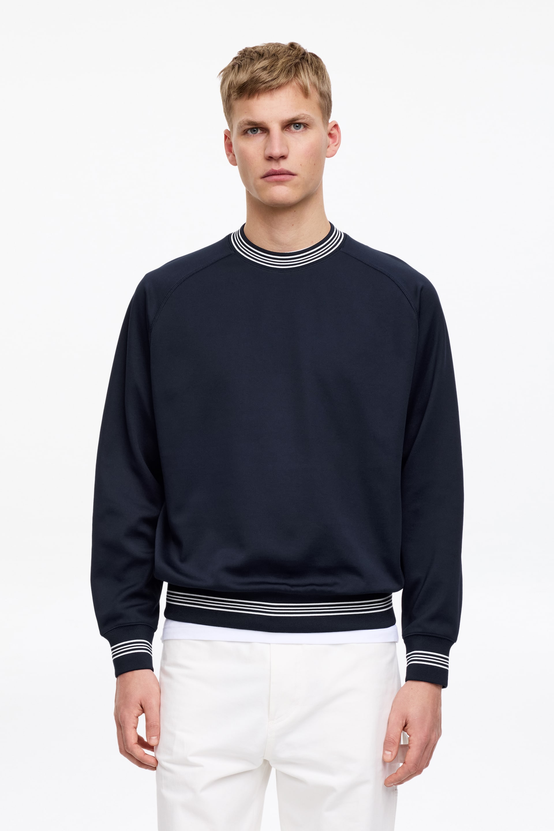 Ingrandisci l'immagine: La modella indossa una felpa blu navy dalla vestibilità ampia, caratterizzata da maniche raglan curve e bordini a costine sullo scollo, sui polsini e sull'orlo inferiore, tutti dettagliati da righe bianche a contrasto. Sotto la felpa si intravede una T-shirt bianca.