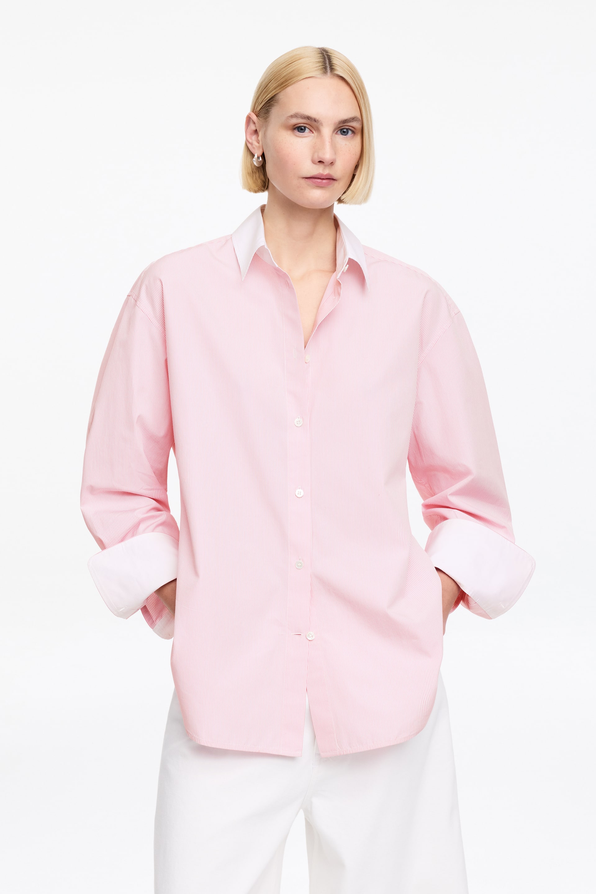 Grotere afbeelding bekijken: Een overhemd met lange mouwen en knoopsluiting, vervaardigd van roze en wit gestreepte katoenpopeline, wordt gedragen door het model. Het heeft een contrasterende witte kraag en witte manchetten, die zijn omgeslagen. Het model staat met de handen in de zakken, waardoor de ontspannen maar geklede silhouet van het overhemd wordt getoond.