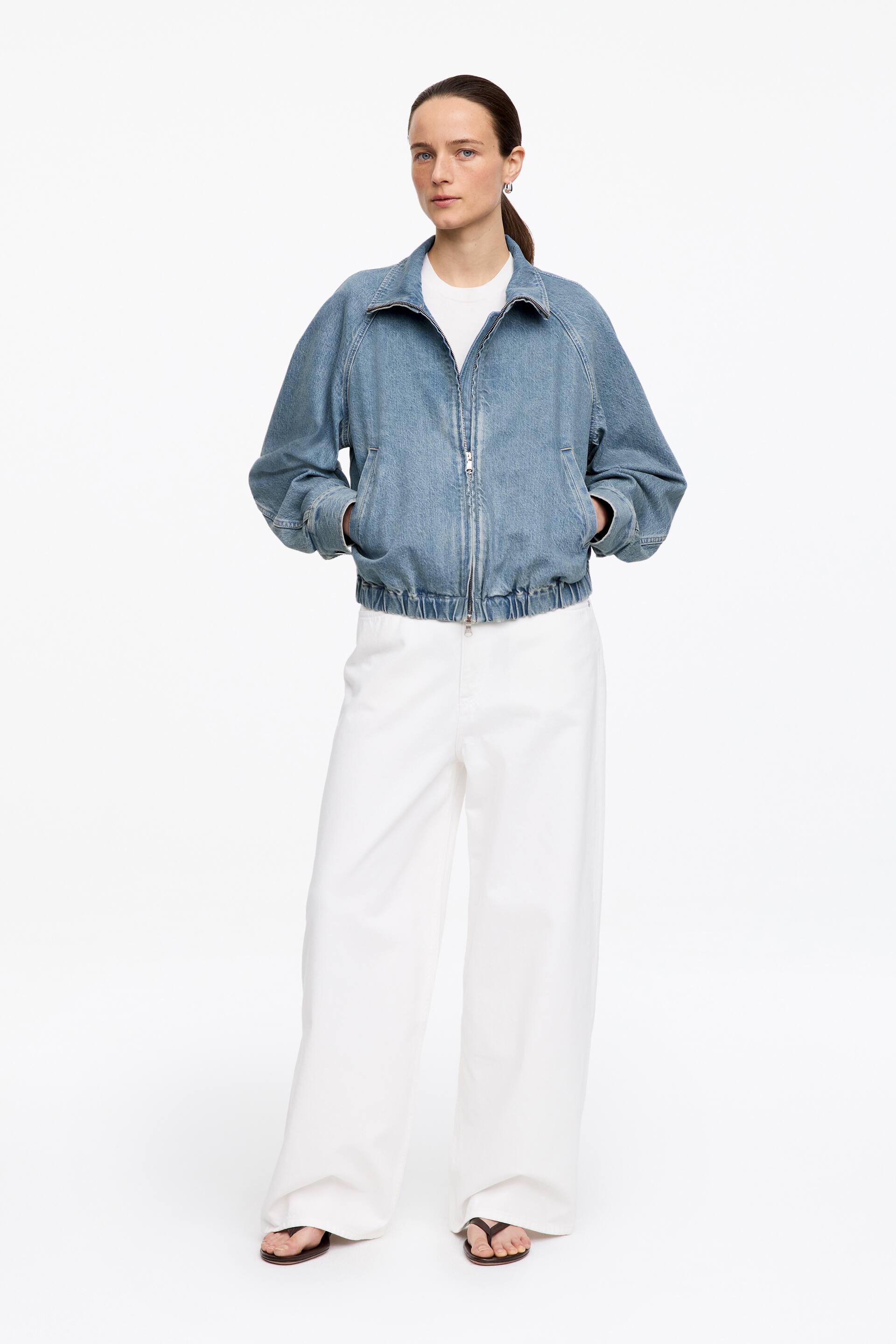 Se større billede: Denimjakke. Denne lyseblå denim bomberjakke har en ståkrave, raglanærmer, en lynlås i fuld længde og to forlommer. Den er designet med elastiske manchetter og en elastisk kant, og den bæres over en hvid t-shirt.