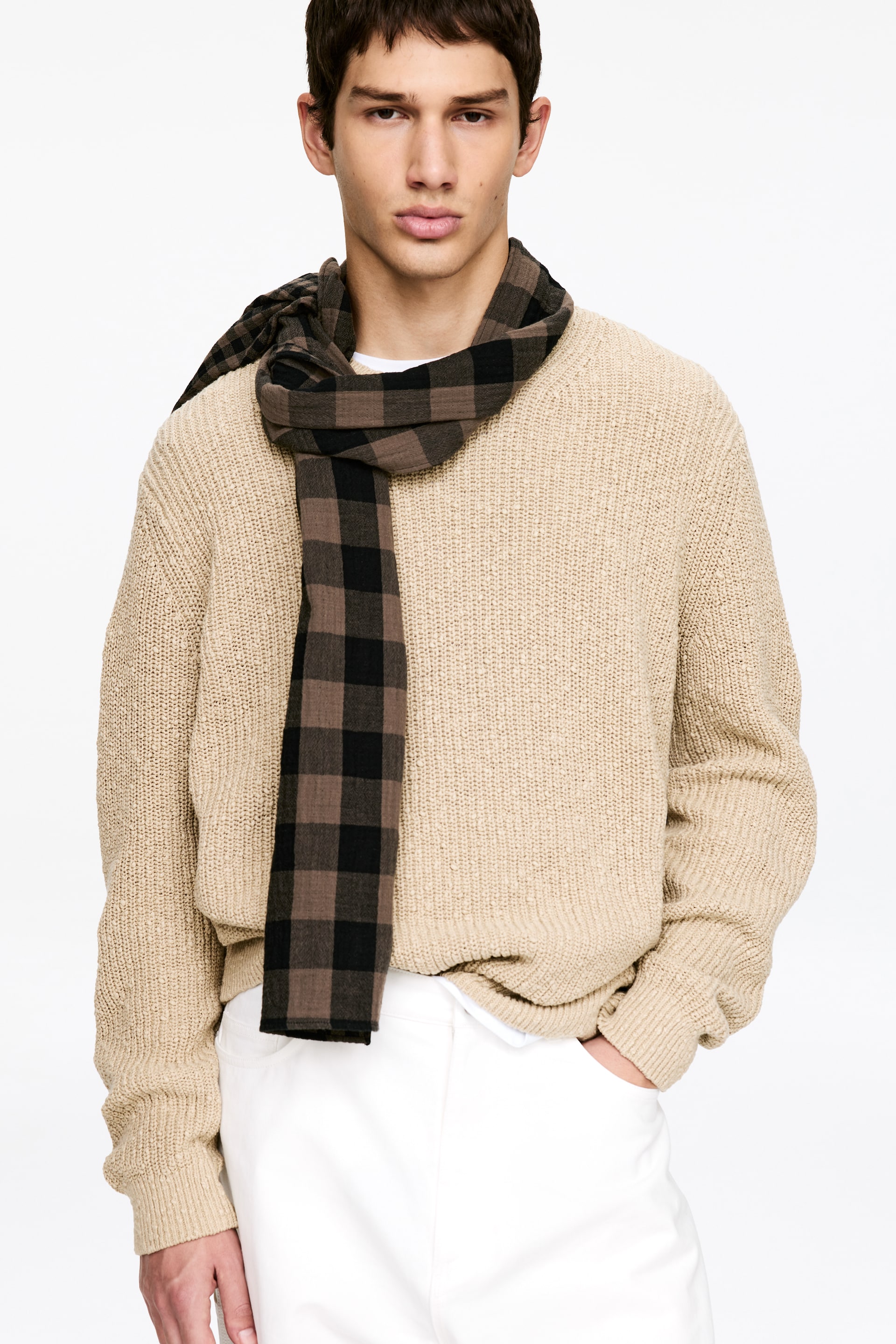 Grotere afbeelding bekijken: De beige gebreide trui toont een gestructureerde, los gebreide constructie met lange mouwen, geribde manchetten en een geribde zoom. Het wordt gepresenteerd vanuit een frontale aanzicht, met een bruin en zwart geruite sjaal om de nek gedrapeerd en een witte onderhemd zichtbaar onder de zoom.