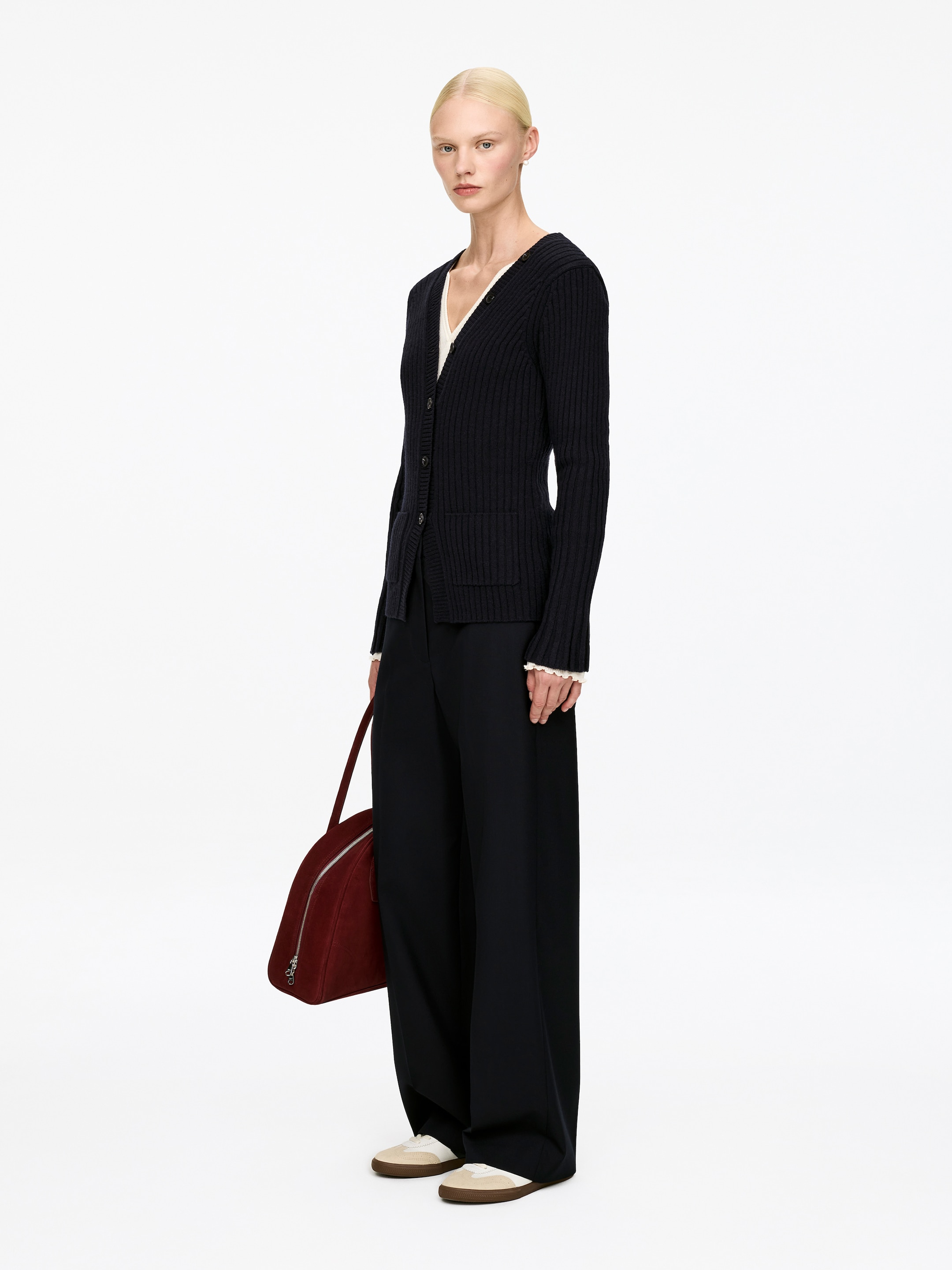 Visualizza immagine più grande: Un cardigan in maglia a coste scuro è presentato da un leggero angolo, caratterizzato da uno scollo a V, chiusura frontale con bottoni, maniche lunghe con polsini a coste e due tasche applicate frontali. Una maglia di colore chiaro è stratificata sotto, con il suo polsino visibile al polso.