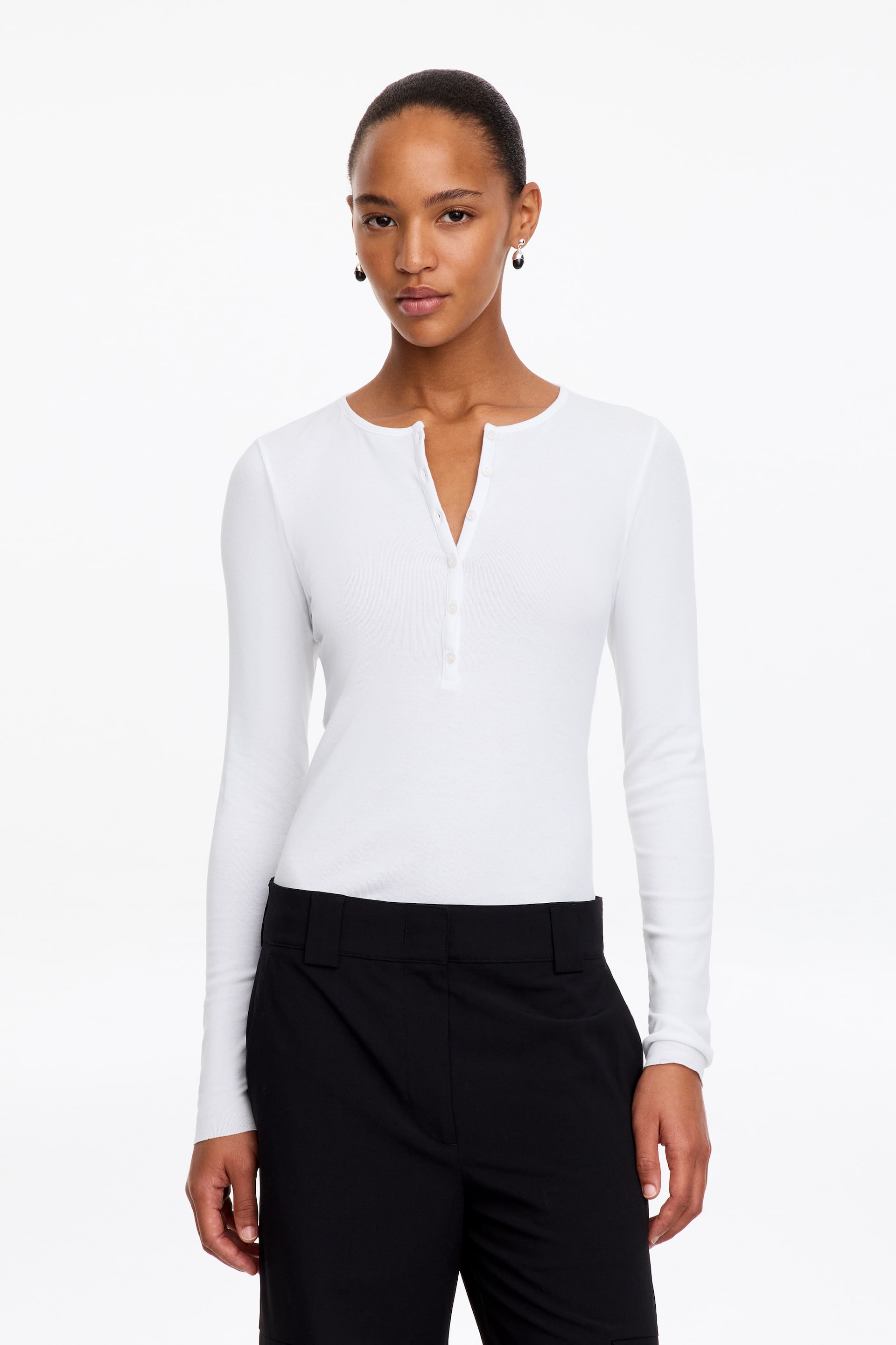 Größeres Bild anzeigen: Das Model trägt ein weißes Henley-Top mit langen Ärmeln und Rippstruktur, mit schmaler Knopfleiste und kleinen Knöpfen, die am Kragen teilweise offen gelassen sind. Das Top ist in eine schwarze, taillierte Hose gesteckt.