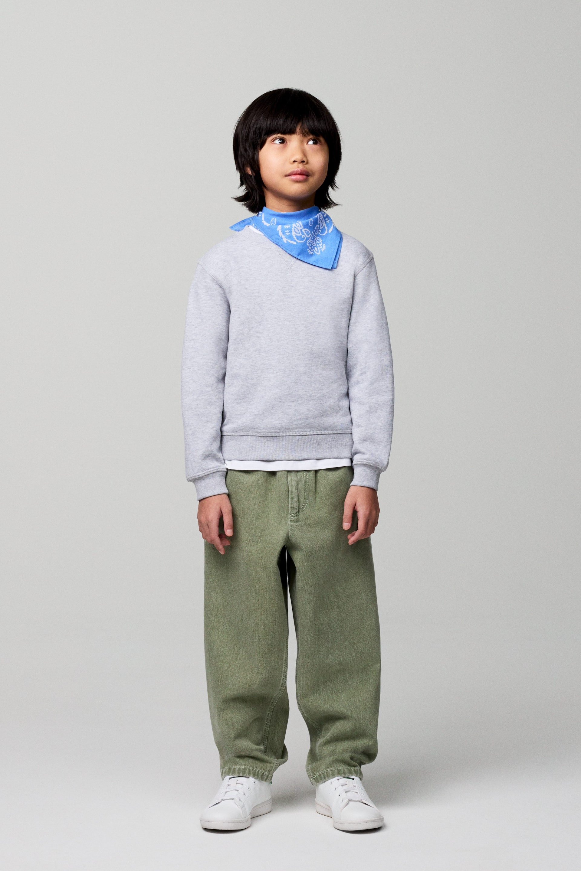 Näytä suurempi kuva: Vaaleanharmaa pyöreäkauluksinen collegepaita, jossa on ribatut hihansuut ja helma, on puettu valkoisen t-paidan päälle ja sinisen valkopaisley-kuvioisen bandanan alle. Väljät oliivinvihreät housut, joissa on joustava vyötärönauha, ovat myös käytössä.