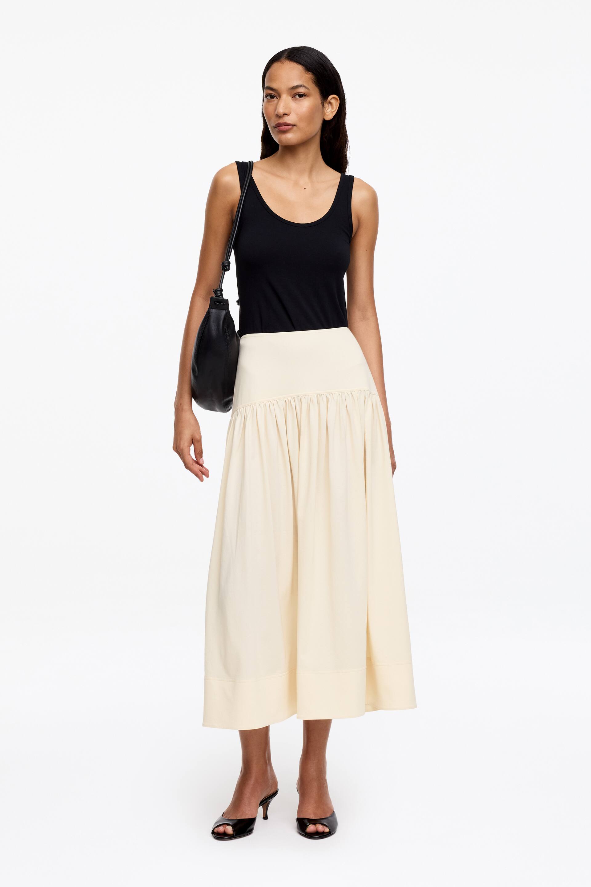 Zobrazit větší obrázek: Černé tílko s kulatým výstřihem je zastrčené do krémové midi sukně s vysokým pasem, která má nabíraný pas a plný, splývavý střih.