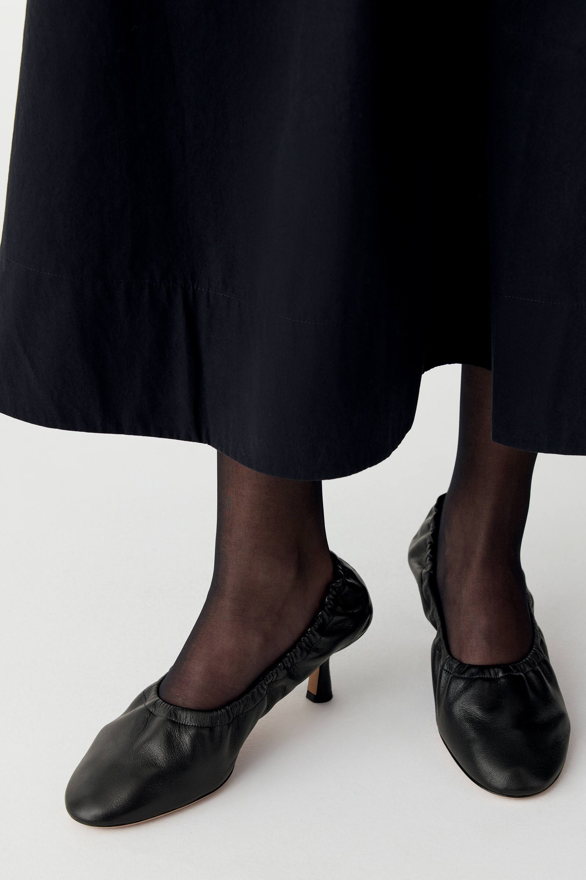 Vis større bilde: Svarte pumps i lær med avrundet tå, rynkedetaljer langs åpningen og en lav kitten heel bæres av modellen. Skoene er kombinert med svarte, transparente strømpebukser og et svart midi-skjørt.