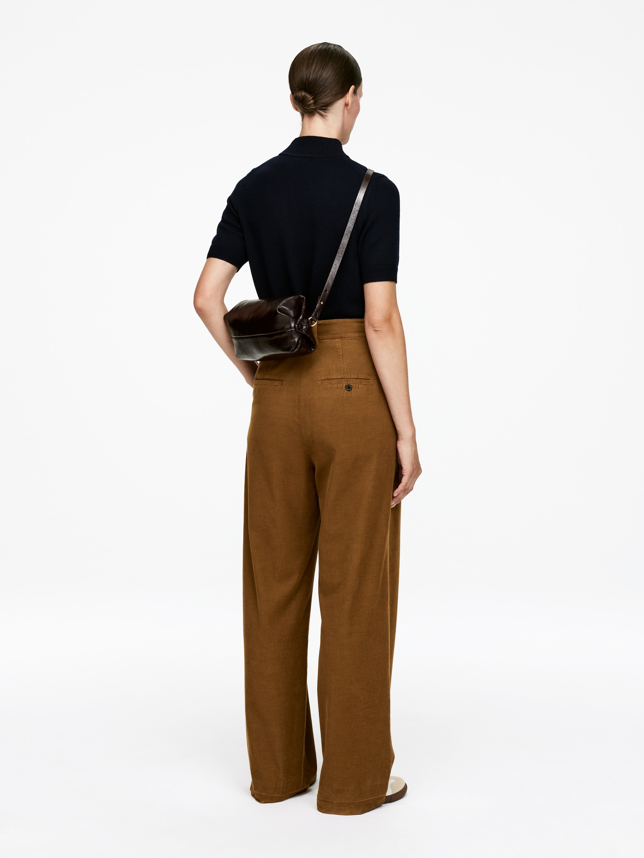 View larger image: A dark brown crossbody bag is being worn, positioned at the wearer's lower back/hip. It is presenting a soft, gathered shape with a smooth finish, and a thin strap is extending diagonally across the back.