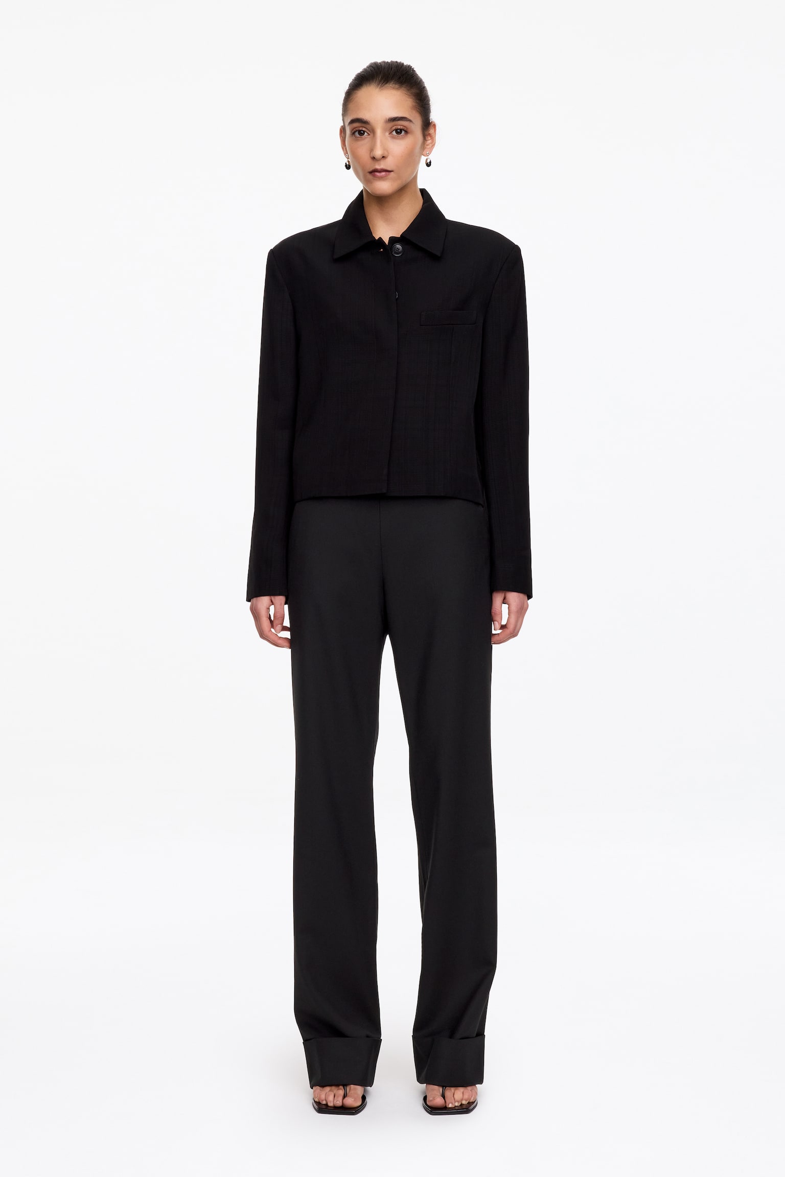 Un pantalon noir est présenté de face, avec une taille haute, un devant épuré et sans couture, et une coupe droite. Le pantalon est terminé par de larges revers pliés aux ourlets. - 1