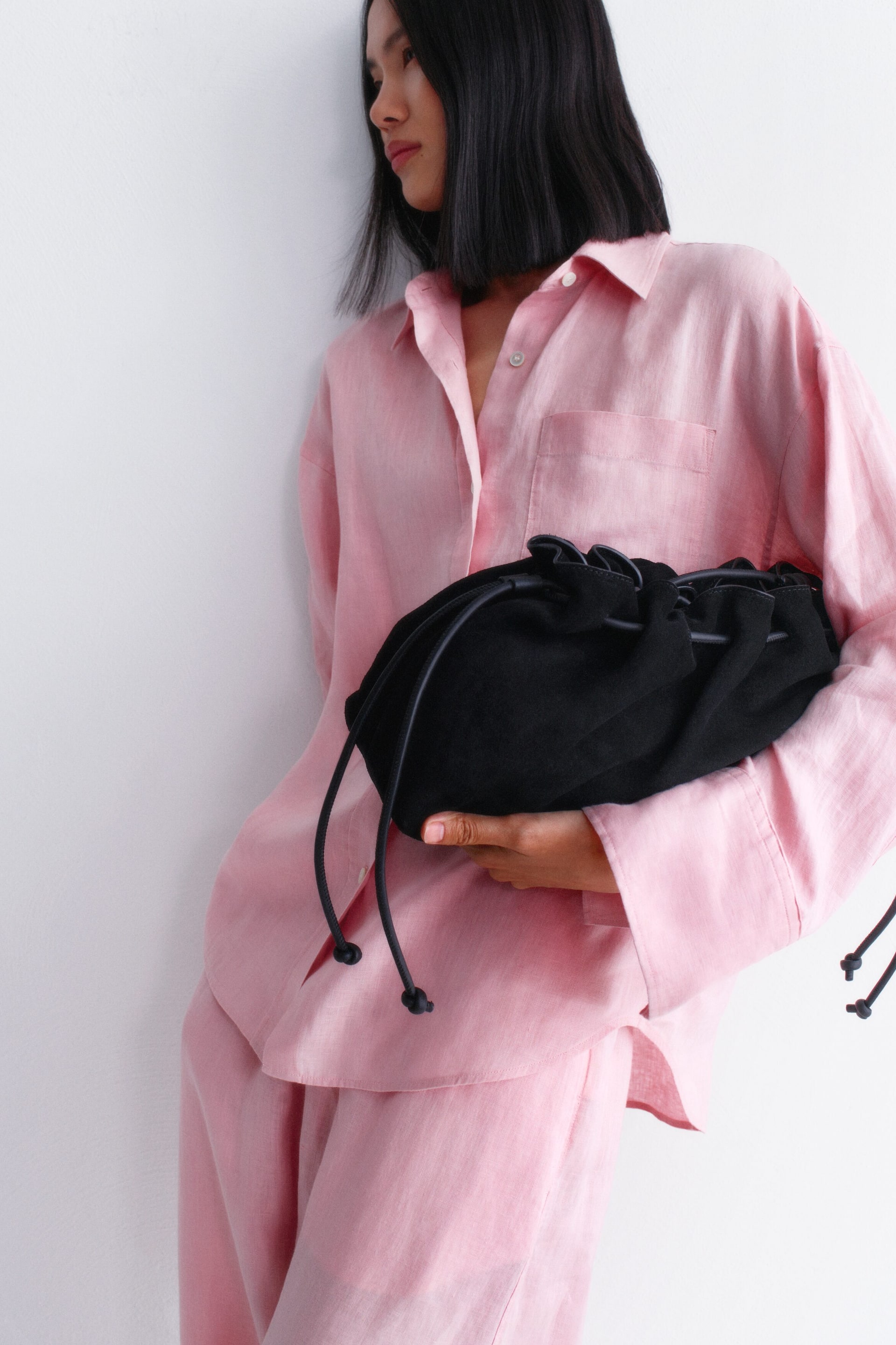 Visualizza immagine più grande: Una borsa a sacchetto nera con coulisse viene tenuta in mano, caratterizzata da una texture morbida e opaca, una parte superiore arricciata e due cordini neri con estremità annodate che pendono dalla parte anteriore.