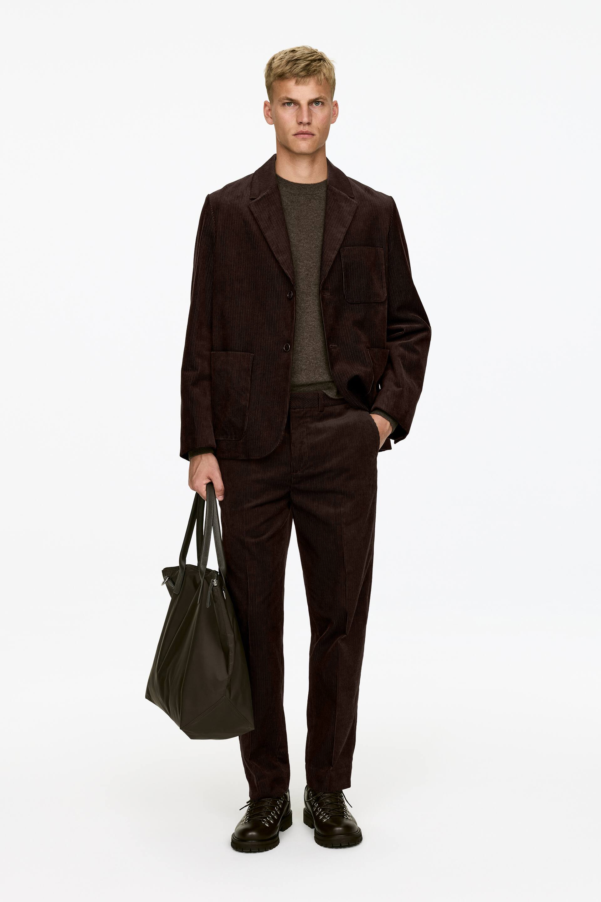 Vis større bilde: Mørkebrune kordfløyelsbukser med vanlig passform, synlige pressfolder og skråstilte frontlommer, bæres av modellen. Modellen står med en hånd i en bukselomme, og buksene styles med en matchende kordfløyelsblazer.